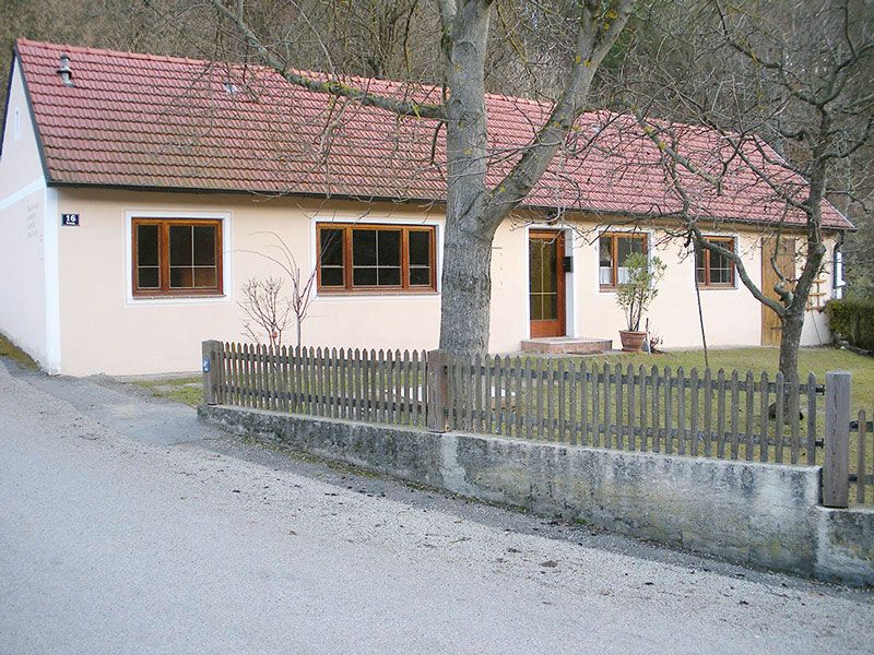 Jednopodlažní rekreační dům s červenou taškovou střechou a světlým nátěrem, obklopený dřevěným plotem a stromy.