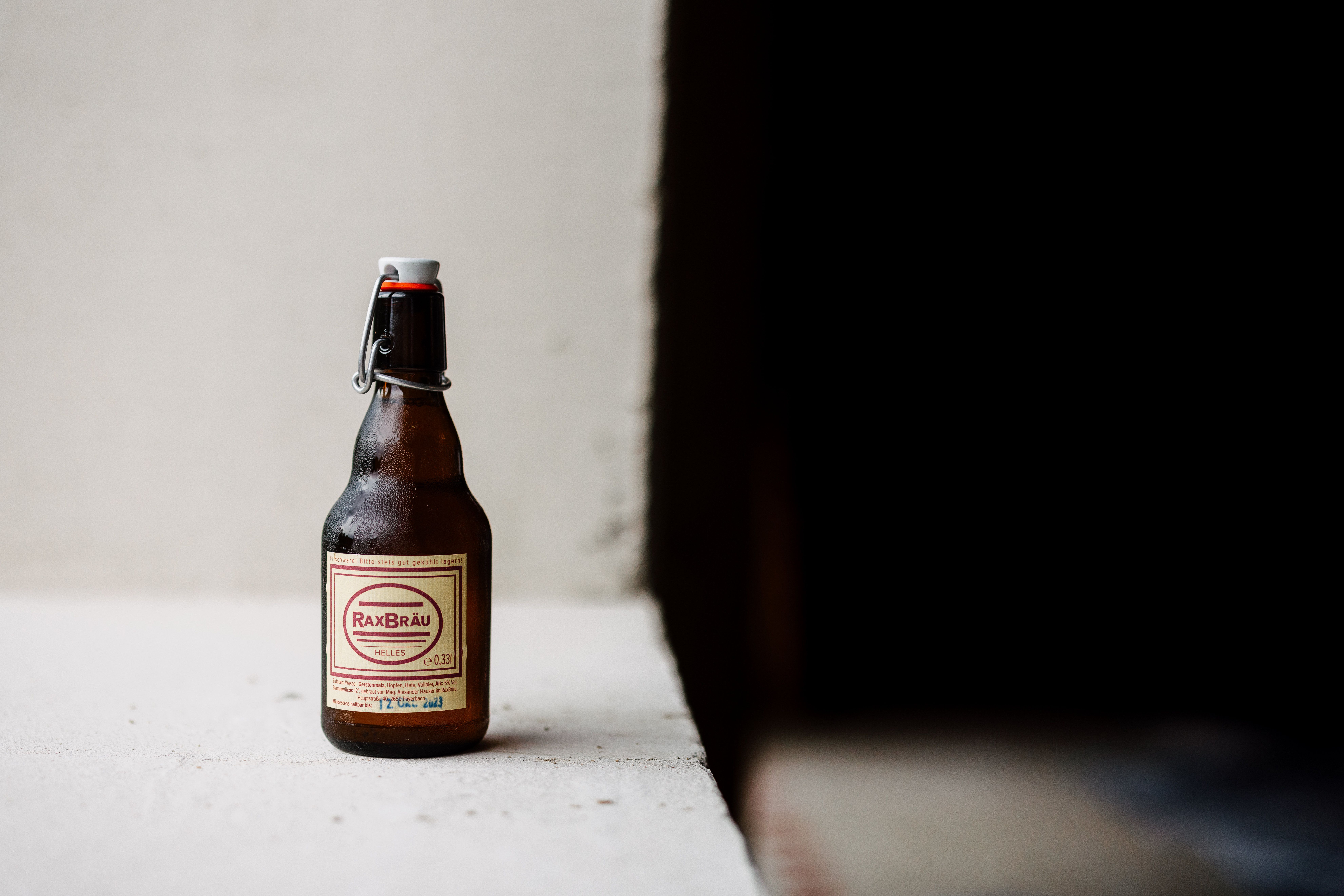 Láhev piva RaxBräu Helles Bier na bílé ploše na tmavém pozadí.