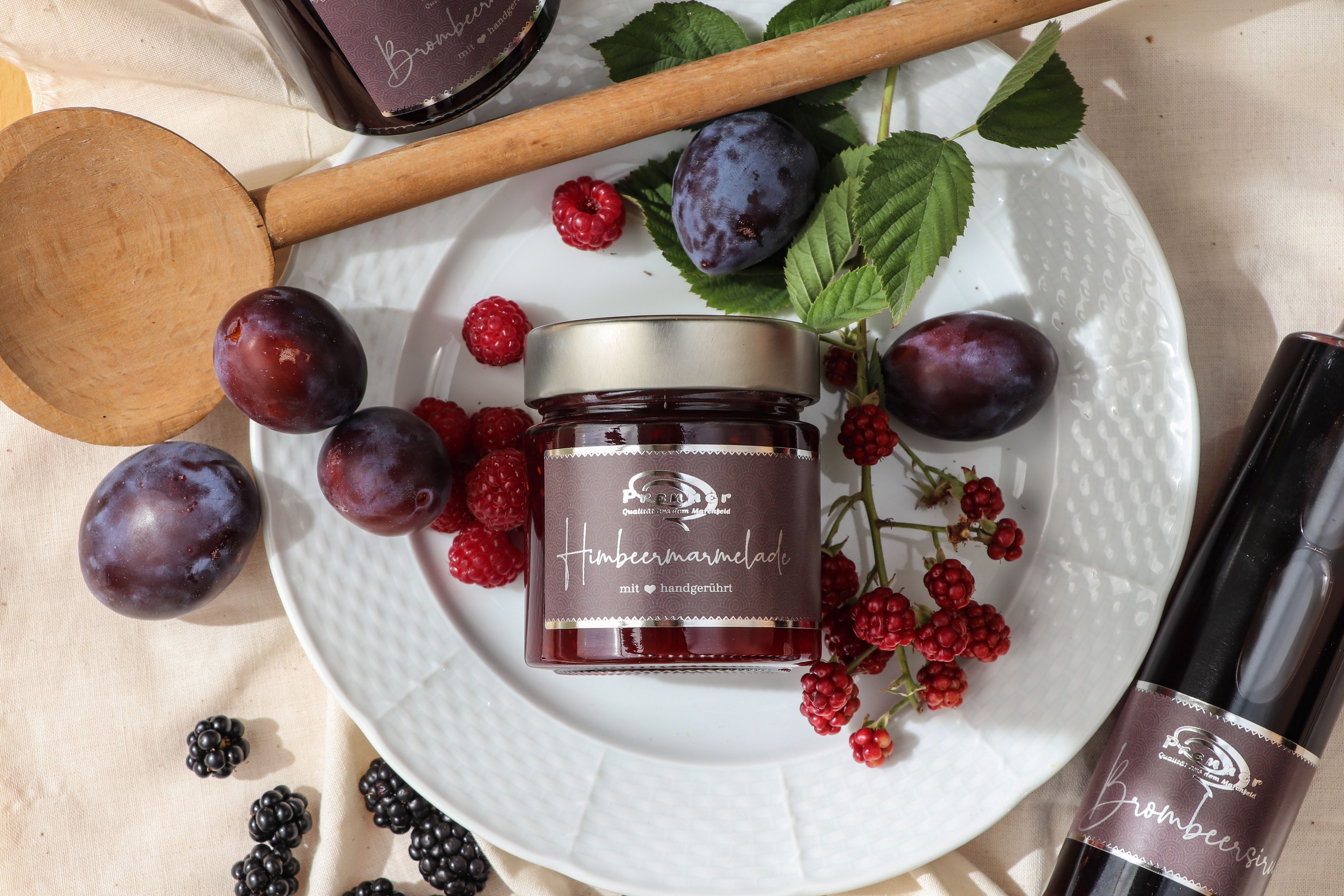 Sklenice malinové marmelády na talíři s čerstvými švestkami, malinami a ostružinami, obklopená dřevěnou lžící a lahví ostružinového sirupu.