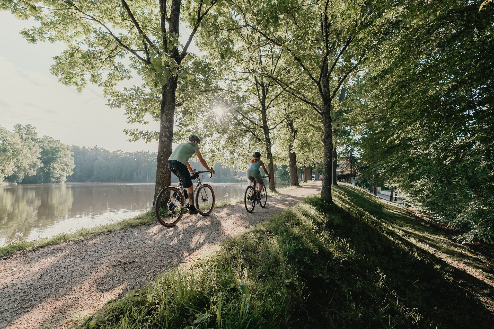 Zwei Personen, die man von hinten sieht, umgeben von Wald und Wasser, die mit ihren Rädern fahren