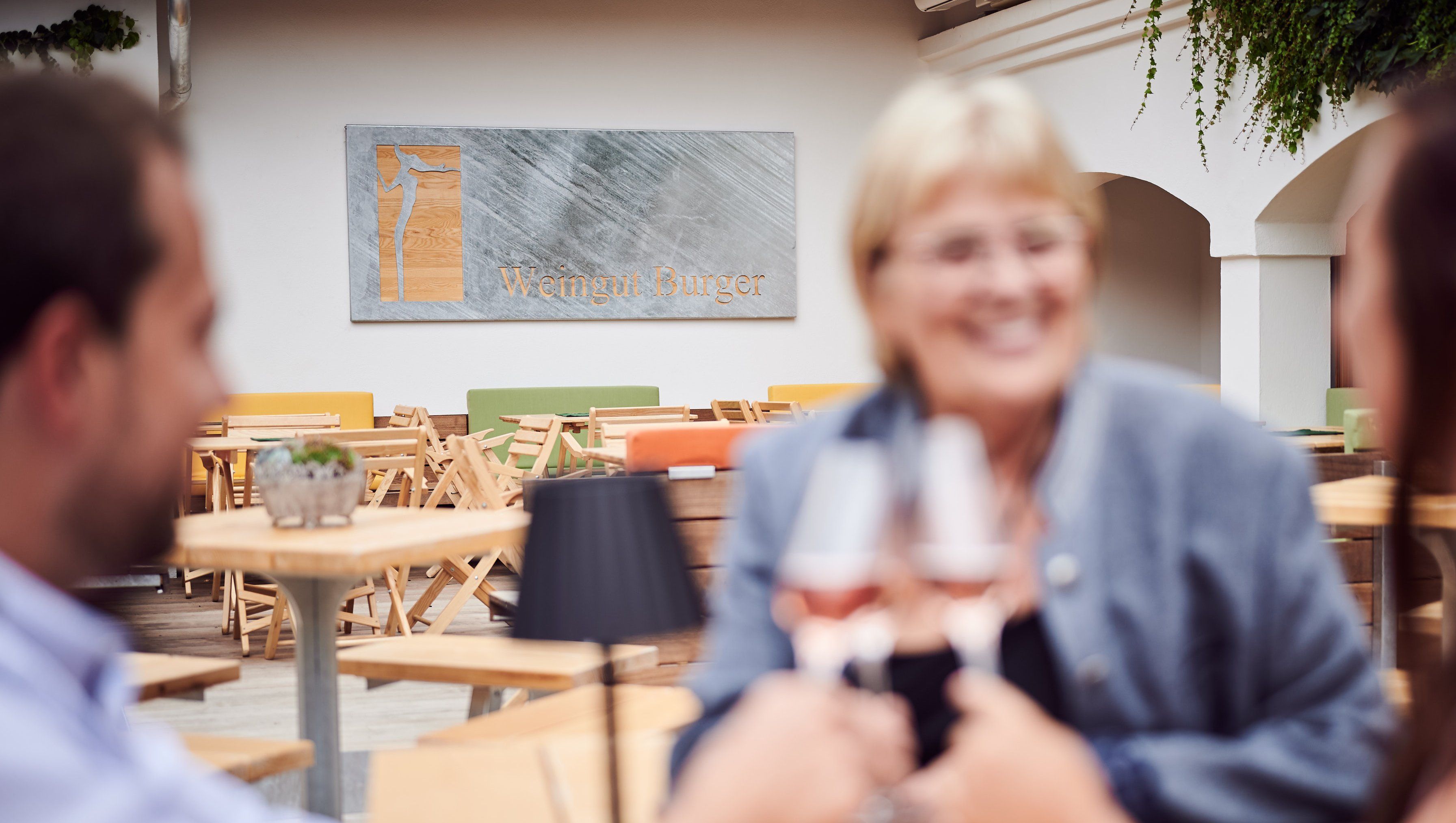 Tři lidé si na dvoře cinknou skleničkami vína a v pozadí je nápis "Weingut Burger".