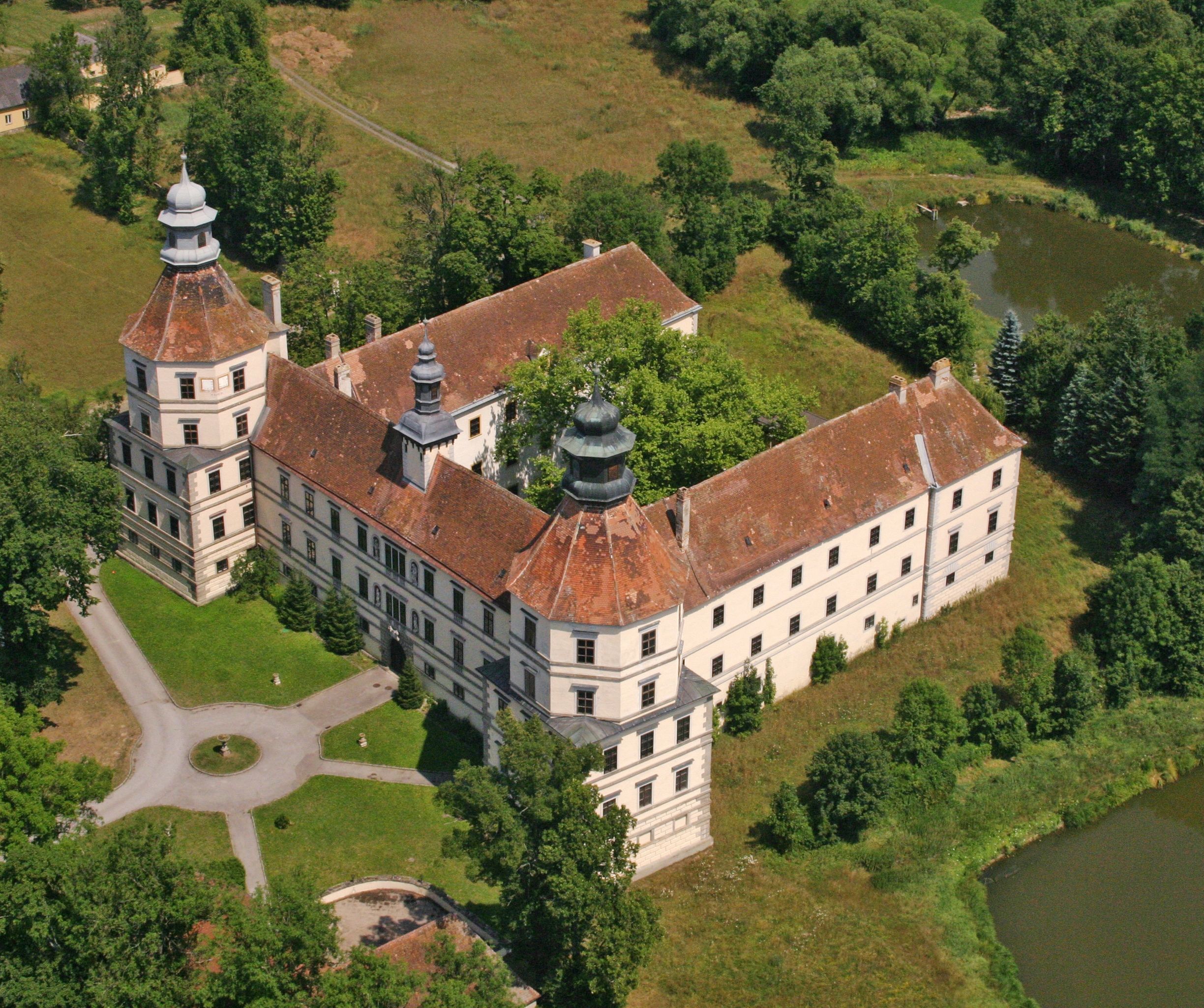 Letecký pohled na historický hrad se dvěma věžemi a vnitřním nádvořím, obklopený stromy a rybníkem.