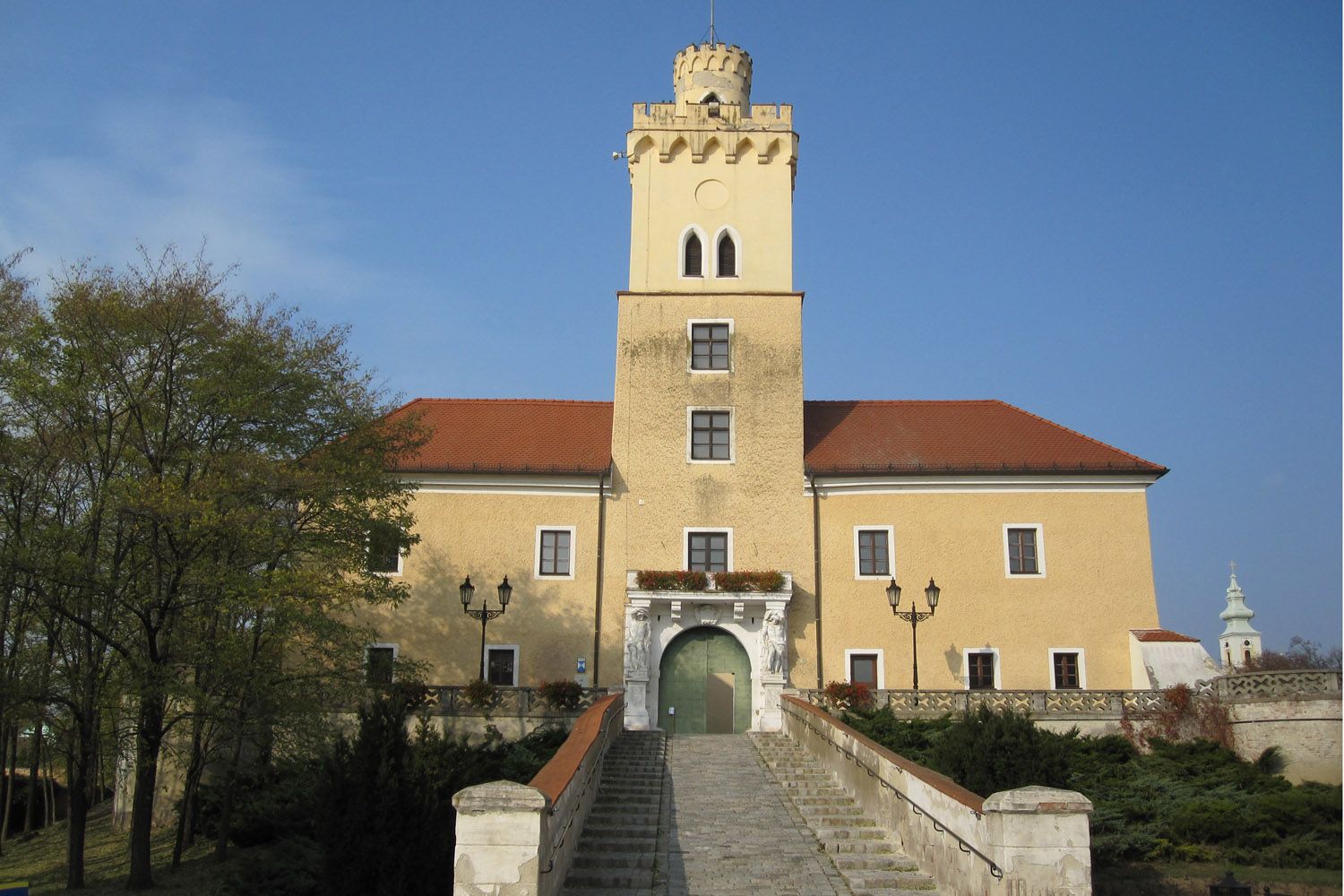 Hrad se žlutou fasádou a červenou střechou, centrální věž, široké kamenné schodiště, modrá obloha.