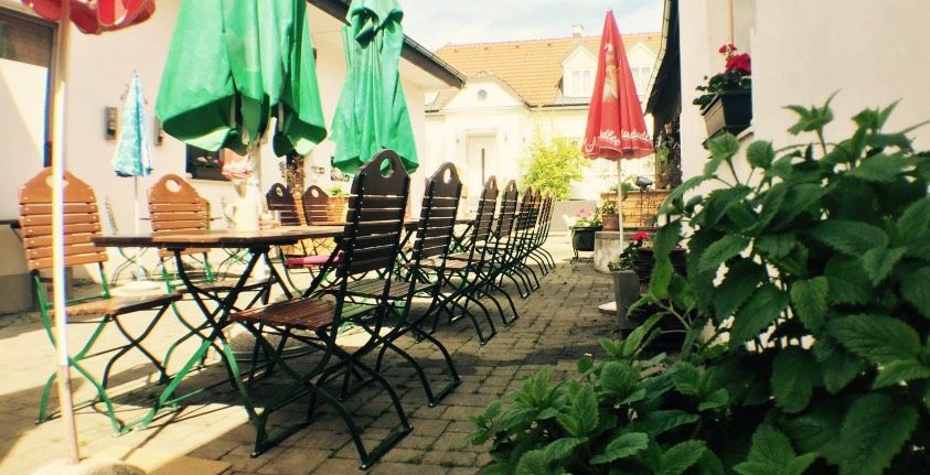 Prázdná zahrada pro hosty s dřevěnými stoly a židlemi, obklopená zelenými slunečníky a rostlinami.