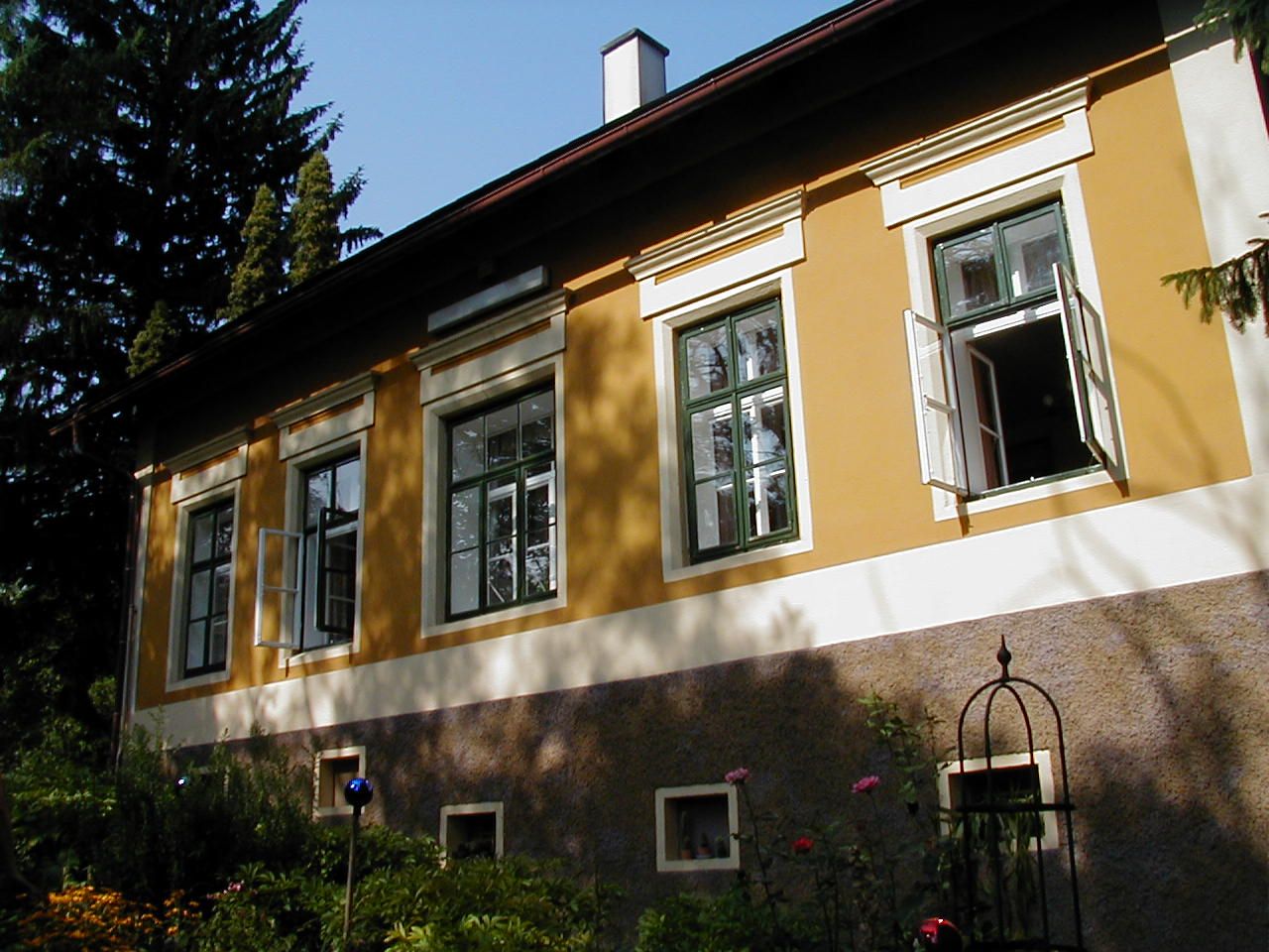 Žlutý dům s otevřenými okny a zahradou v popředí.