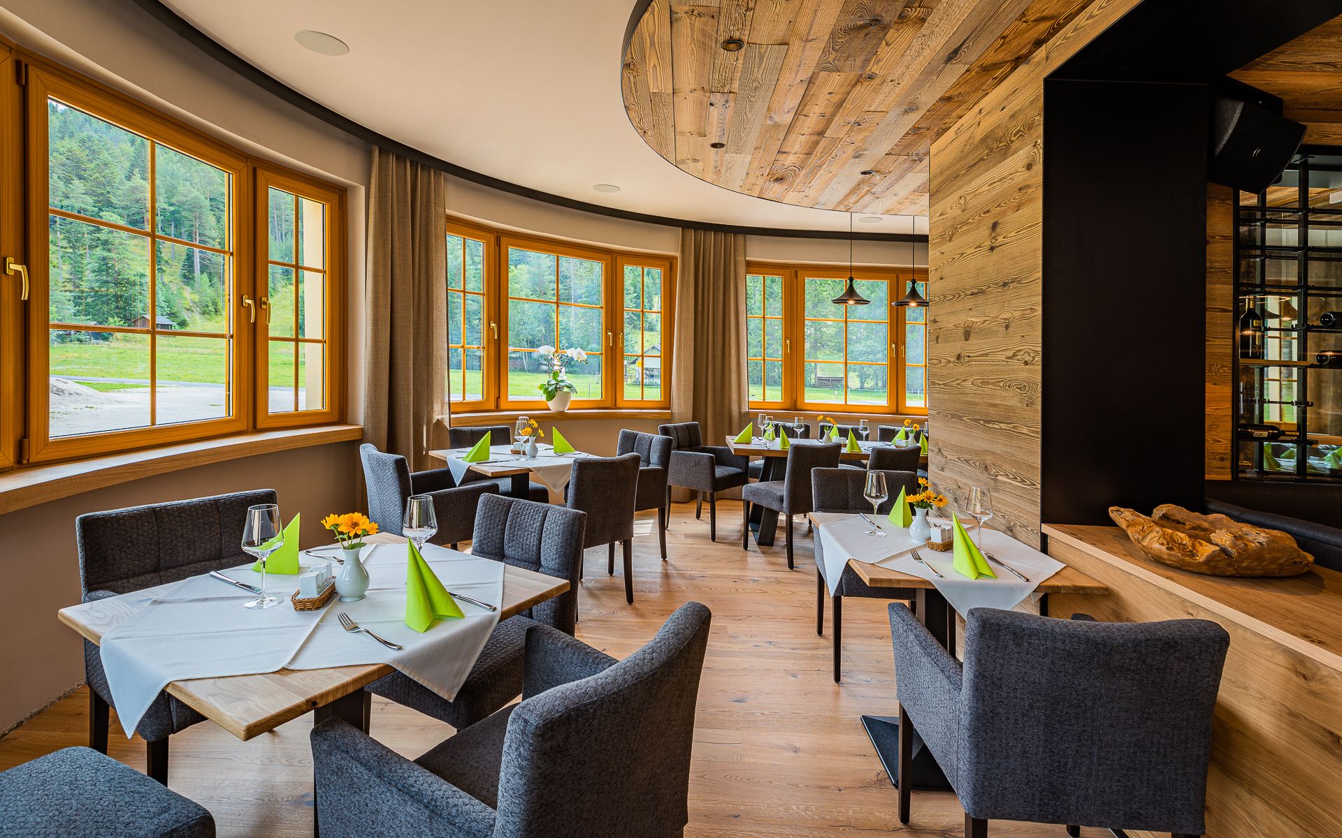 Elegantní restaurace s dřevěnými prvky, prostřenými stoly a velkými okny s výhledem do zeleně.