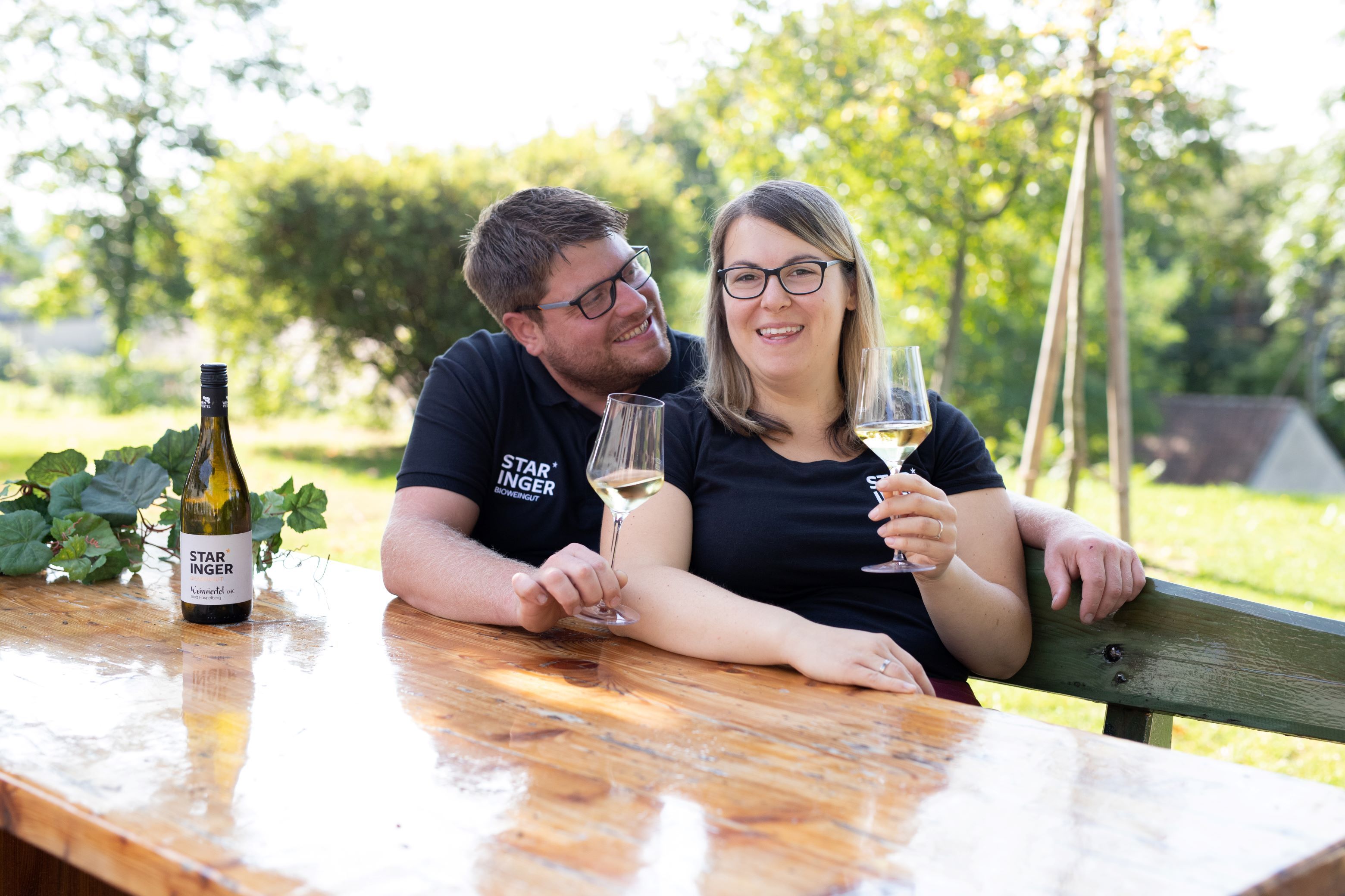 Venku sedí u dřevěného stolu pár se sklenicemi vína. Na stole leží láhev vína s nápisem "Staringer".