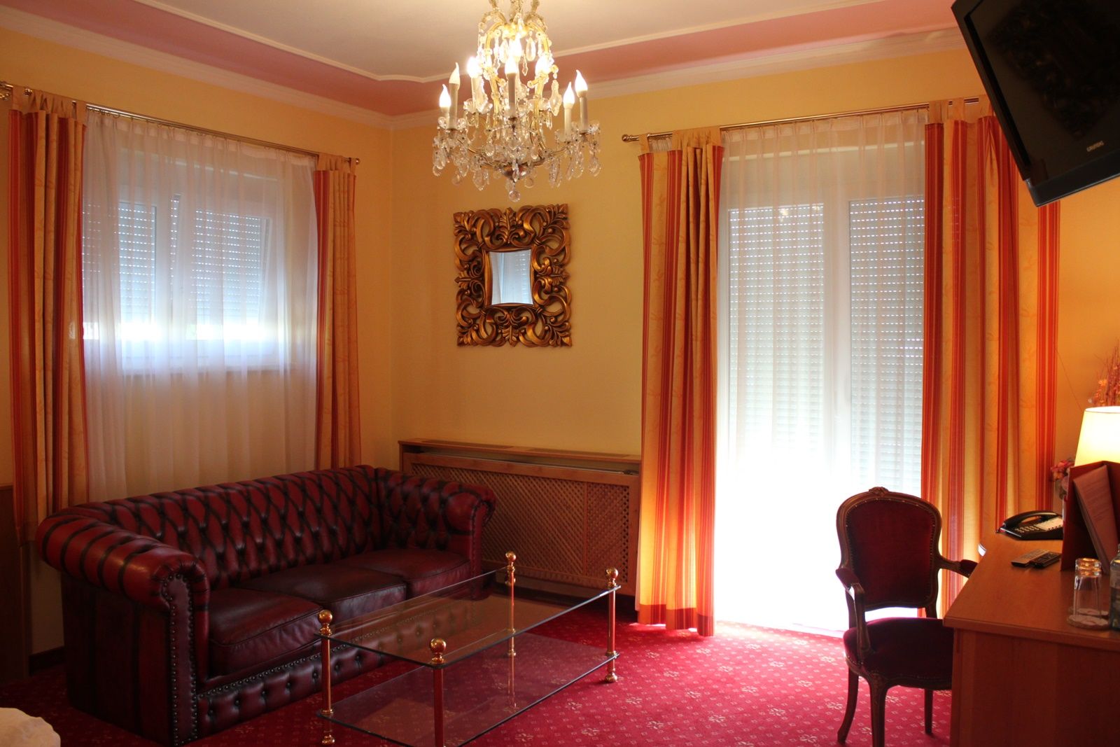 Elegantní obývací pokoj s koženou pohovkou, skleněným stolem, lustrem a červenými závěsy.
