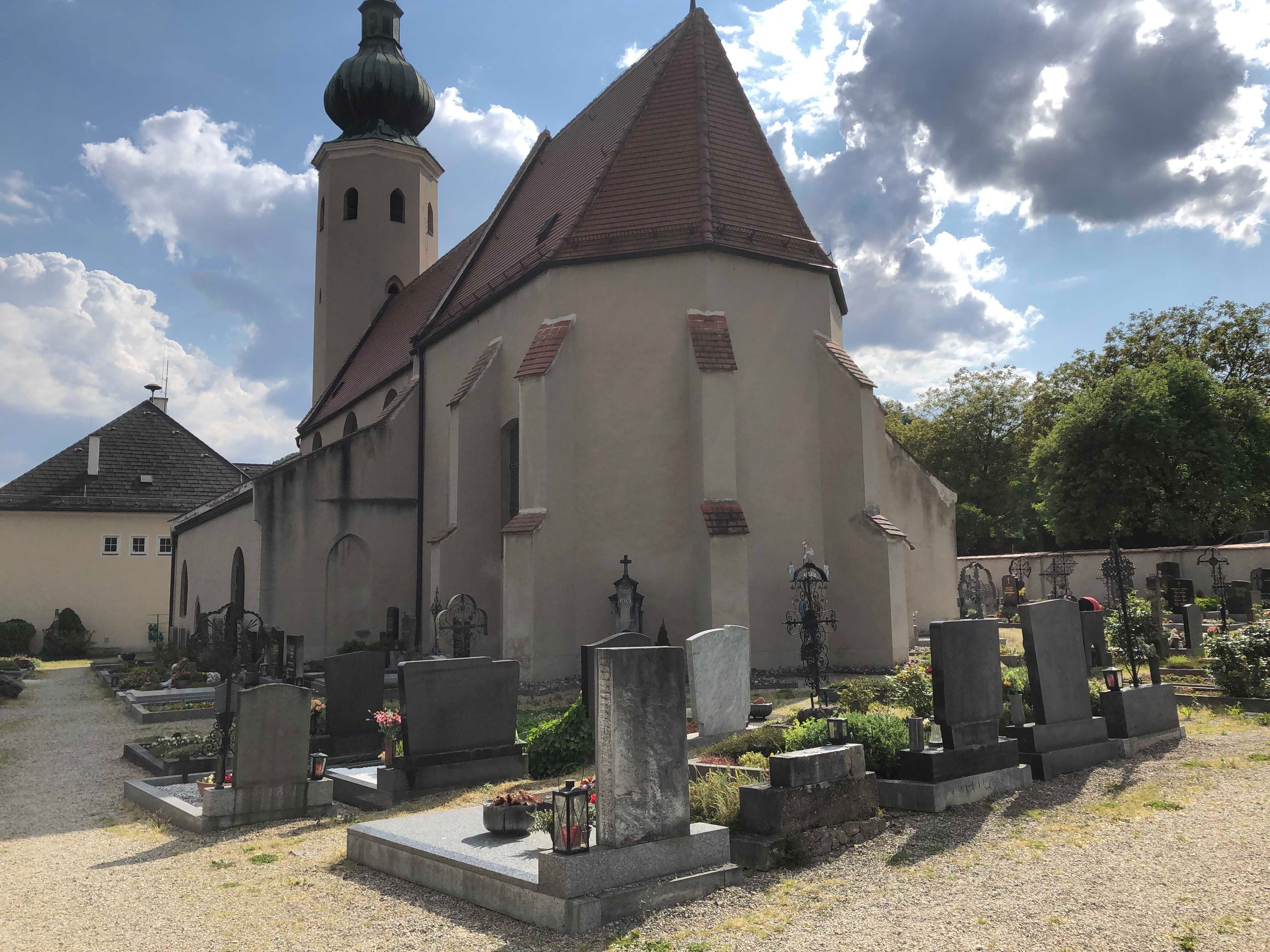 Kostel se hřbitovem a náhrobky v popředí, zatažená obloha.