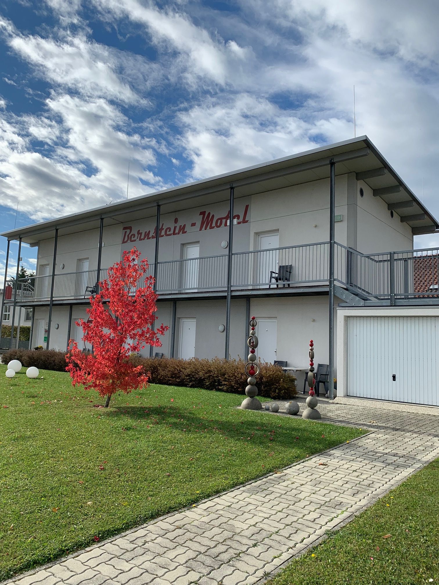 Dvoupatrová budova s nápisem "Bernstein Motel", červený strom v popředí, modrá obloha s mraky.