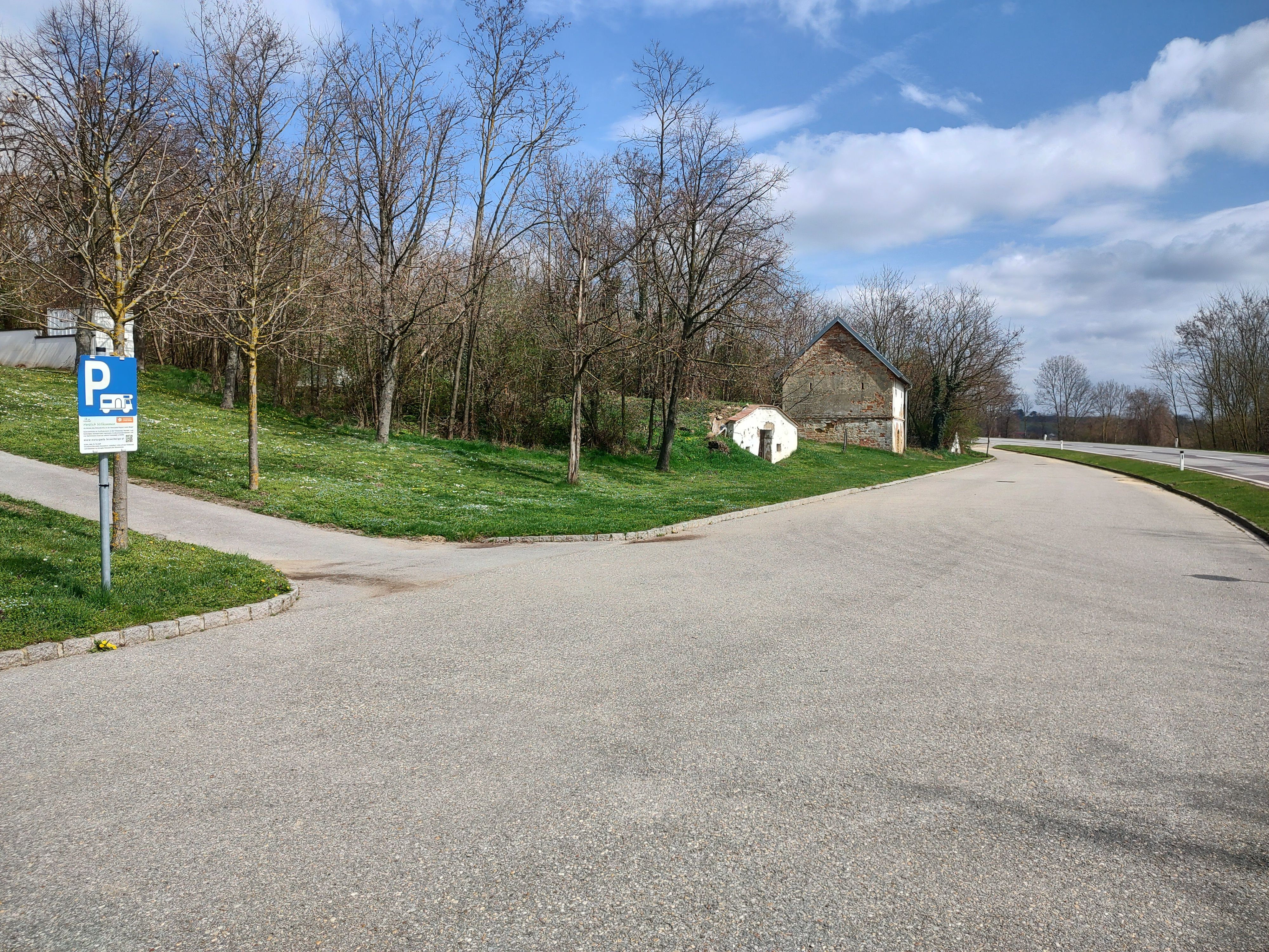 Parkoviště se značkou pro parkování obytných vozů, stromy a stará budova v pozadí.