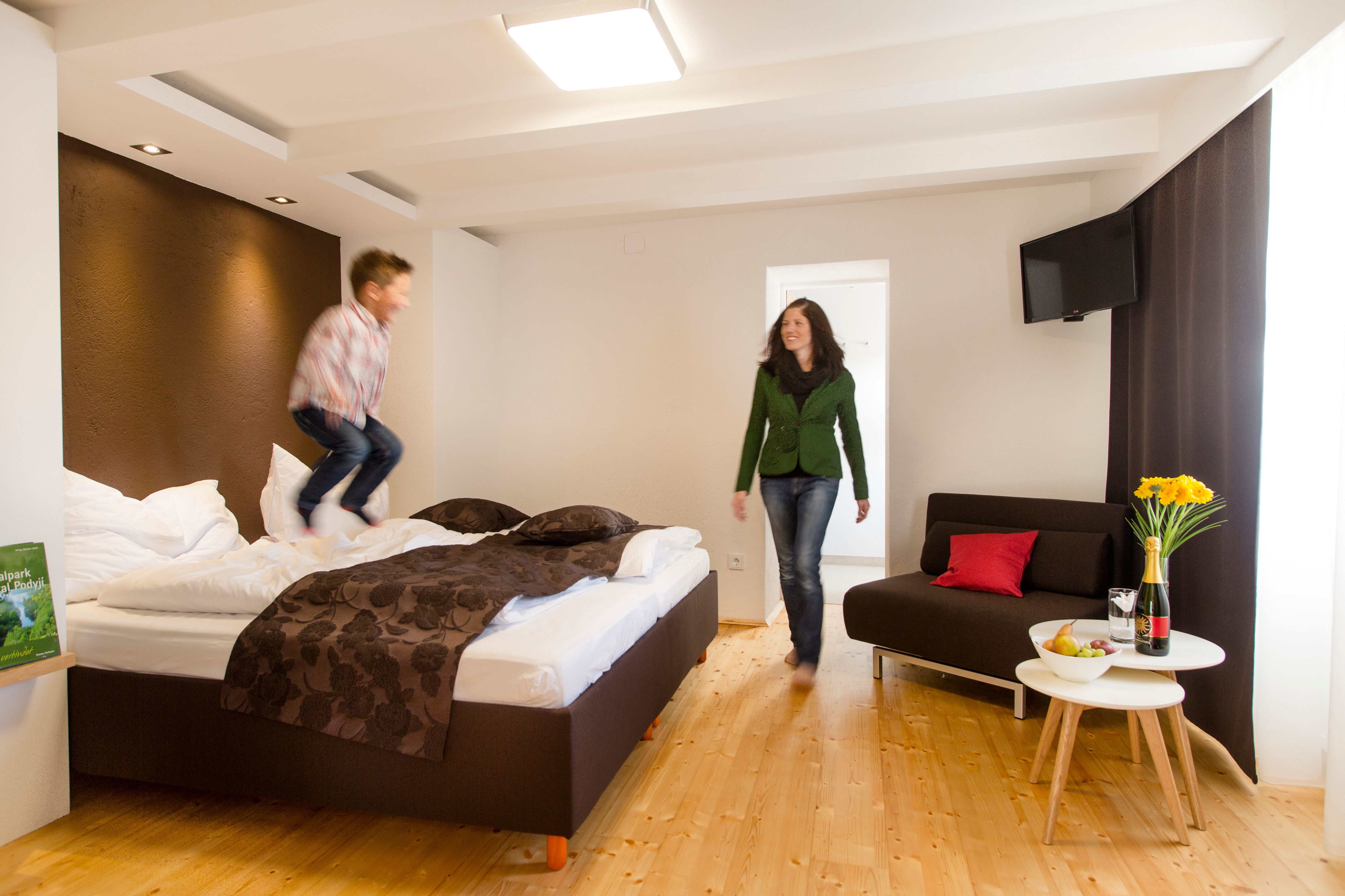 Dítě skáče na posteli v moderním hotelovém pokoji a žena se usmívá. Vedle stojí stůl s květinami a vínem.