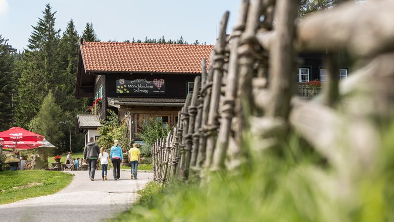 Tři lidé jdou k horskému hostinci, který je obklopen dřevěným plotem a stromy.