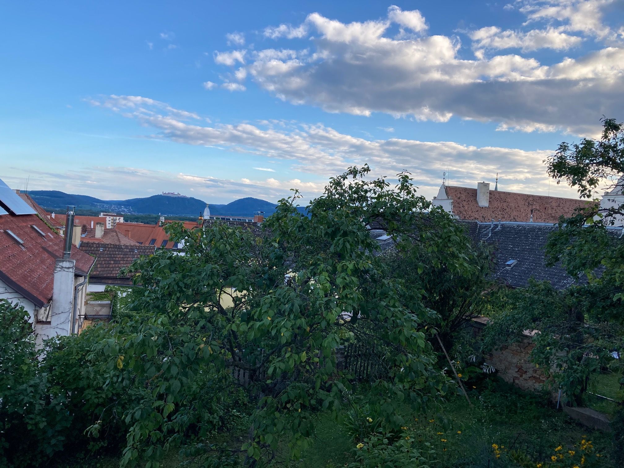 Pohled přes střechy a stromy na kopce a modrou oblohu s mraky.