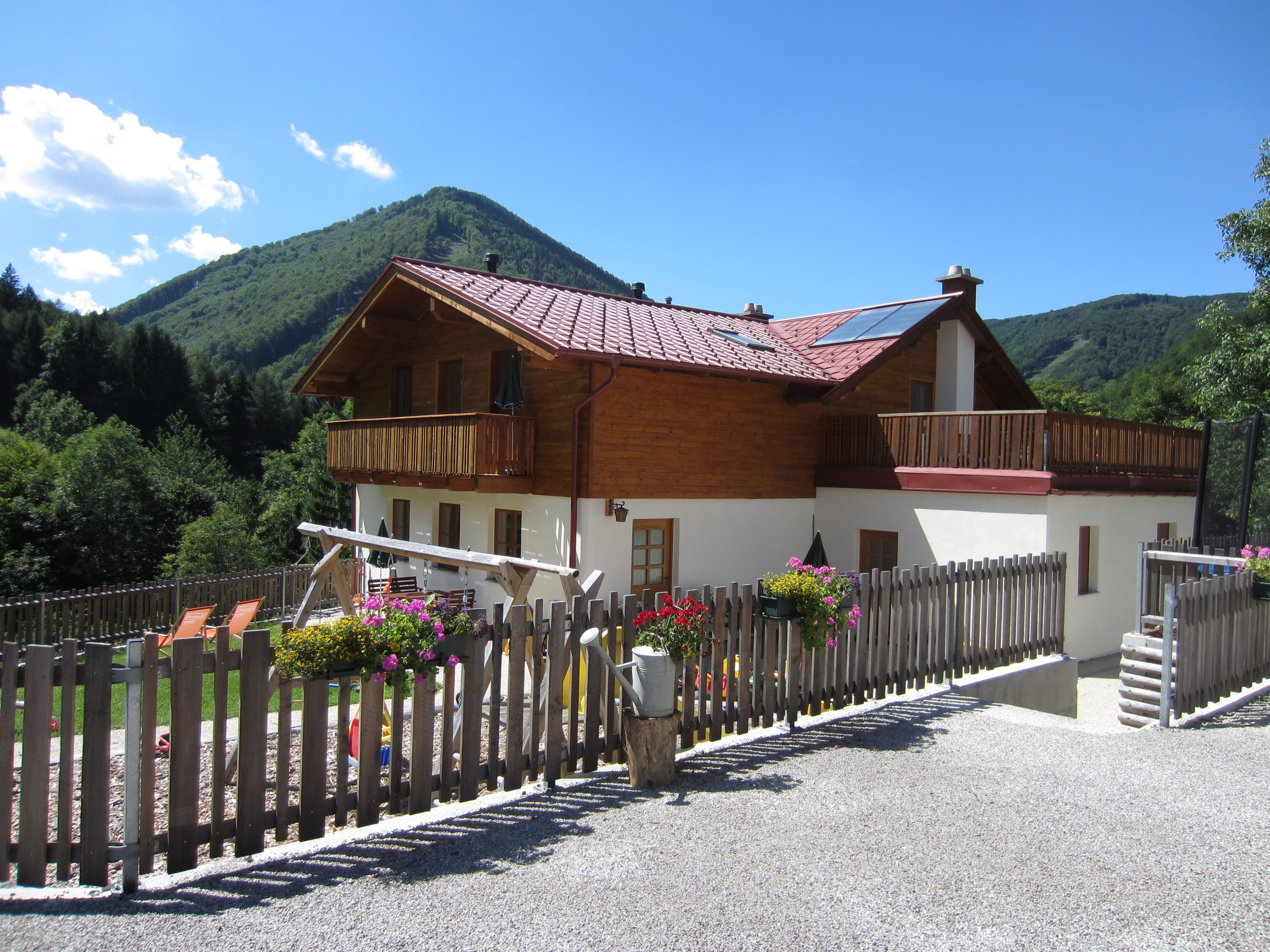 Dvoupodlažní dům s dřevěným balkonem a zahradou, obklopený horami a stromy.