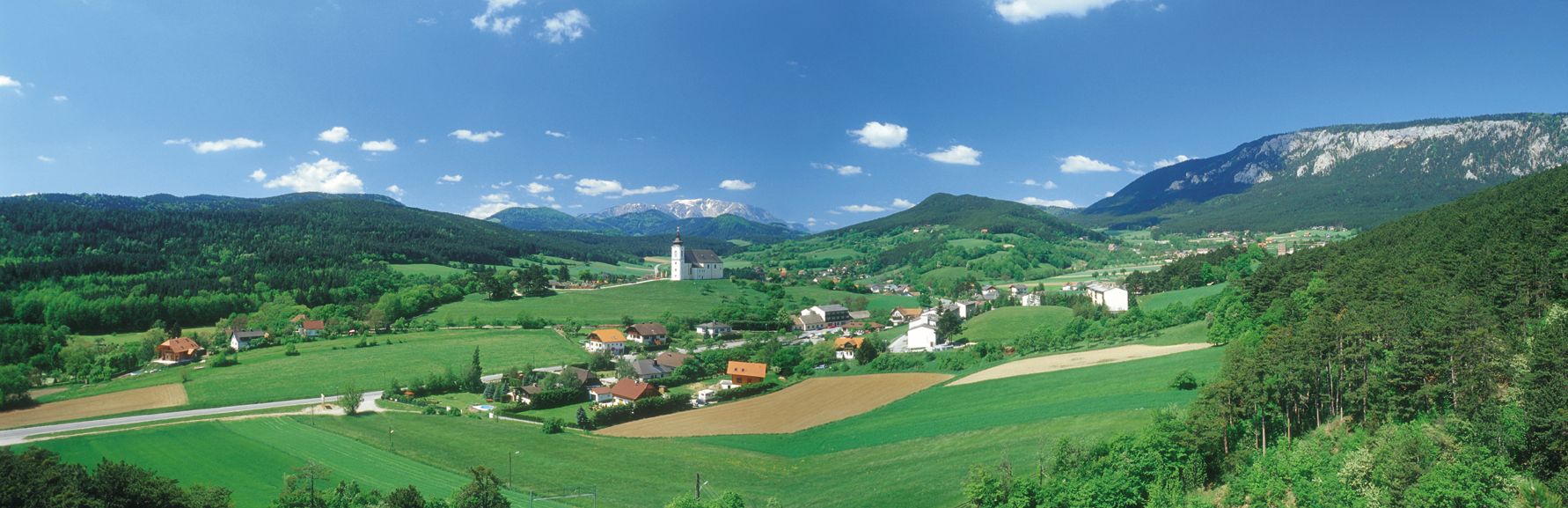 Panoramatický pohled na Höflein s kostelem, zelenými poli a horami v pozadí.