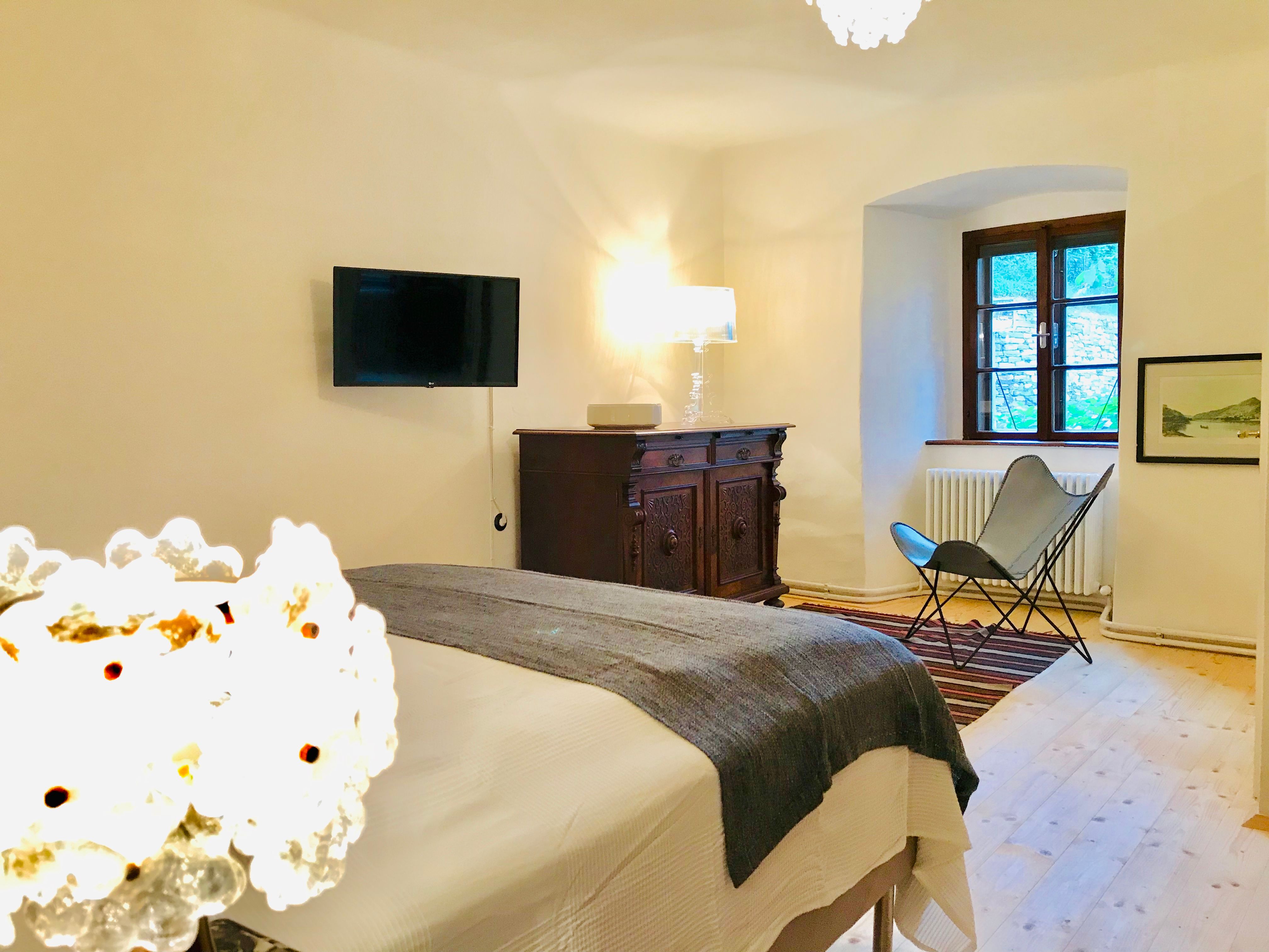 Útulná ložnice s postelí, starožitnou komodou, křeslem a nástěnnou TV.