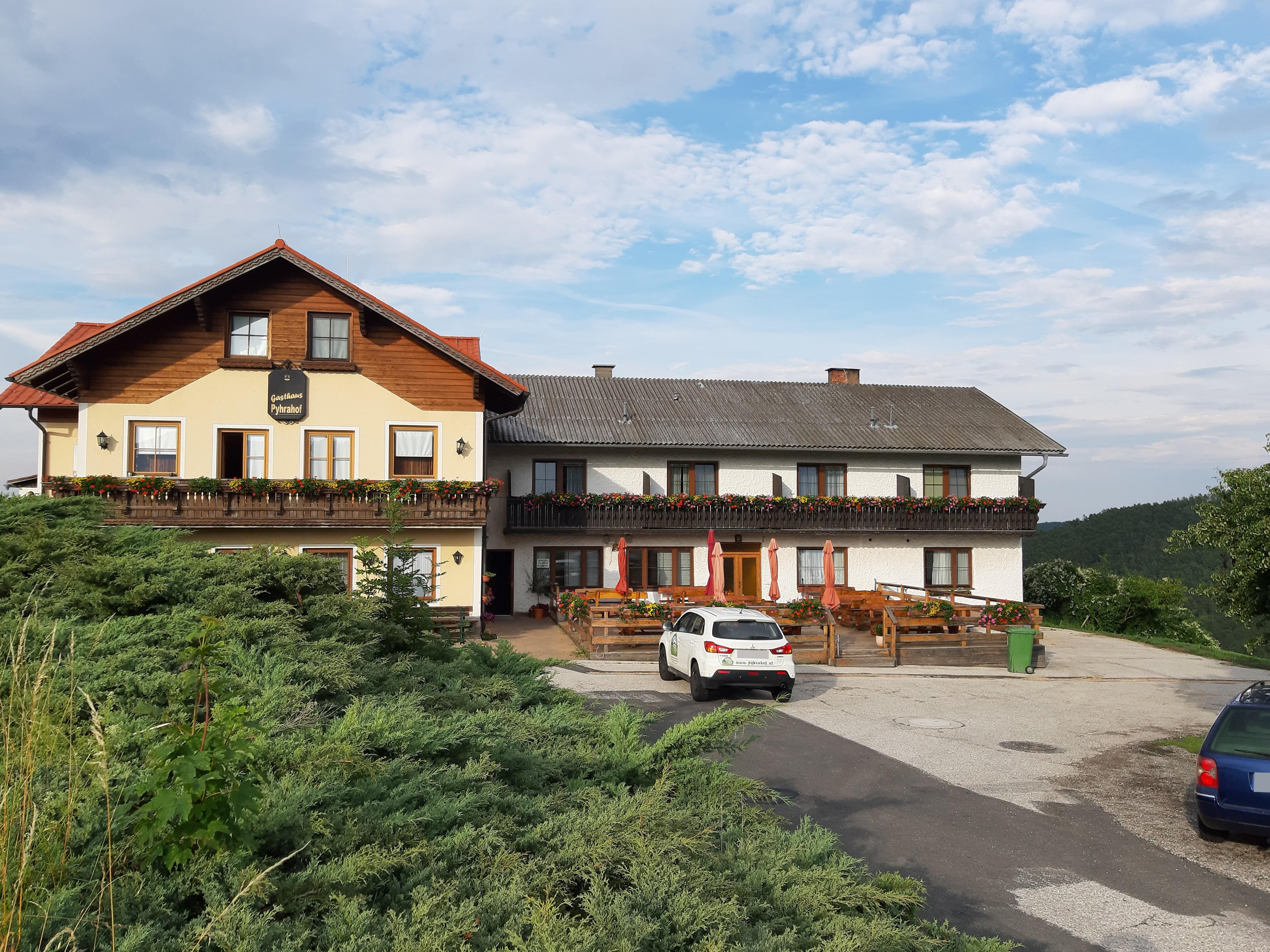 Tradiční hostinec s balkonem a květinovými truhlíky, parkoviště s auty před domem, obklopený zelenou krajinou a modrou oblohou.