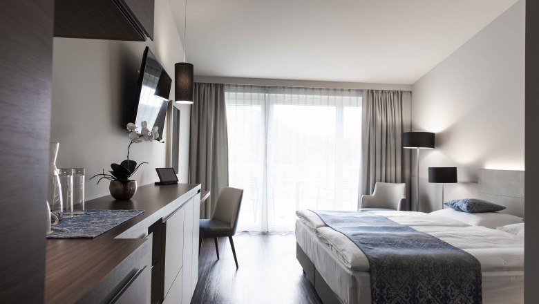 Modern&iacute; hotelov&yacute; pokoj s manželskou postel&iacute;, TV, psac&iacute;m stolem a velk&yacute;mi okny se z&aacute;věsy.