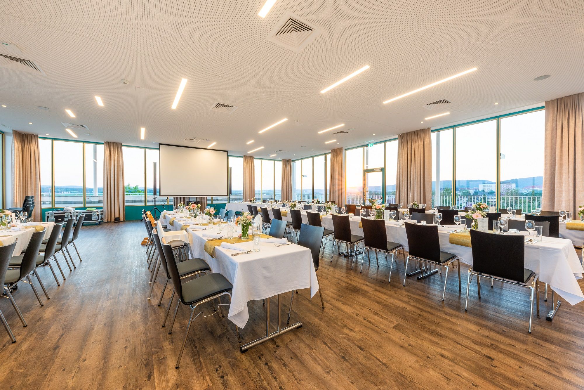 Velká, světlá místnost s krytými stoly a židlemi, velkými okny a projektorem.