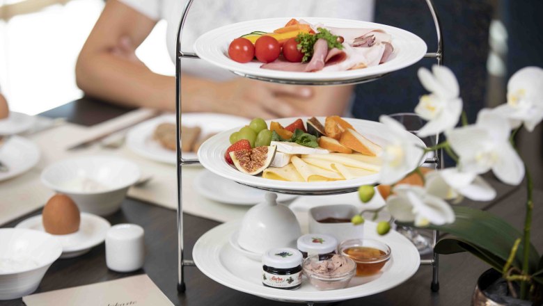 Sn&iacute;daňov&yacute; stůl s et&aacute;ž&iacute; plnou s&yacute;rů, uzenin, ovoce a pomaz&aacute;nek. V popřed&iacute; b&iacute;l&aacute; orchidej.