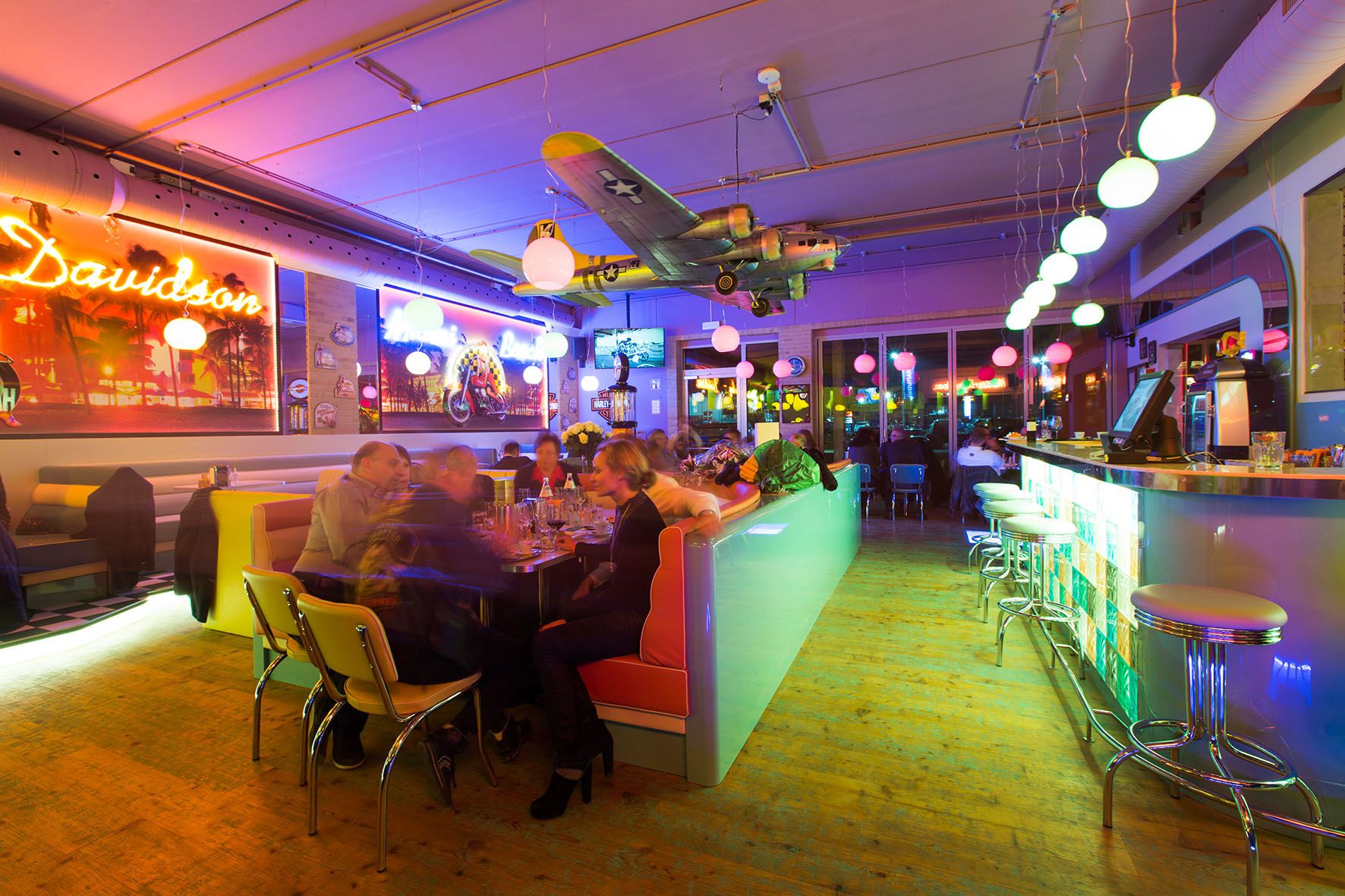 Pohled do interiéru barevně osvětlené restaurace s barem, stoly a modelem letadla na stropě.