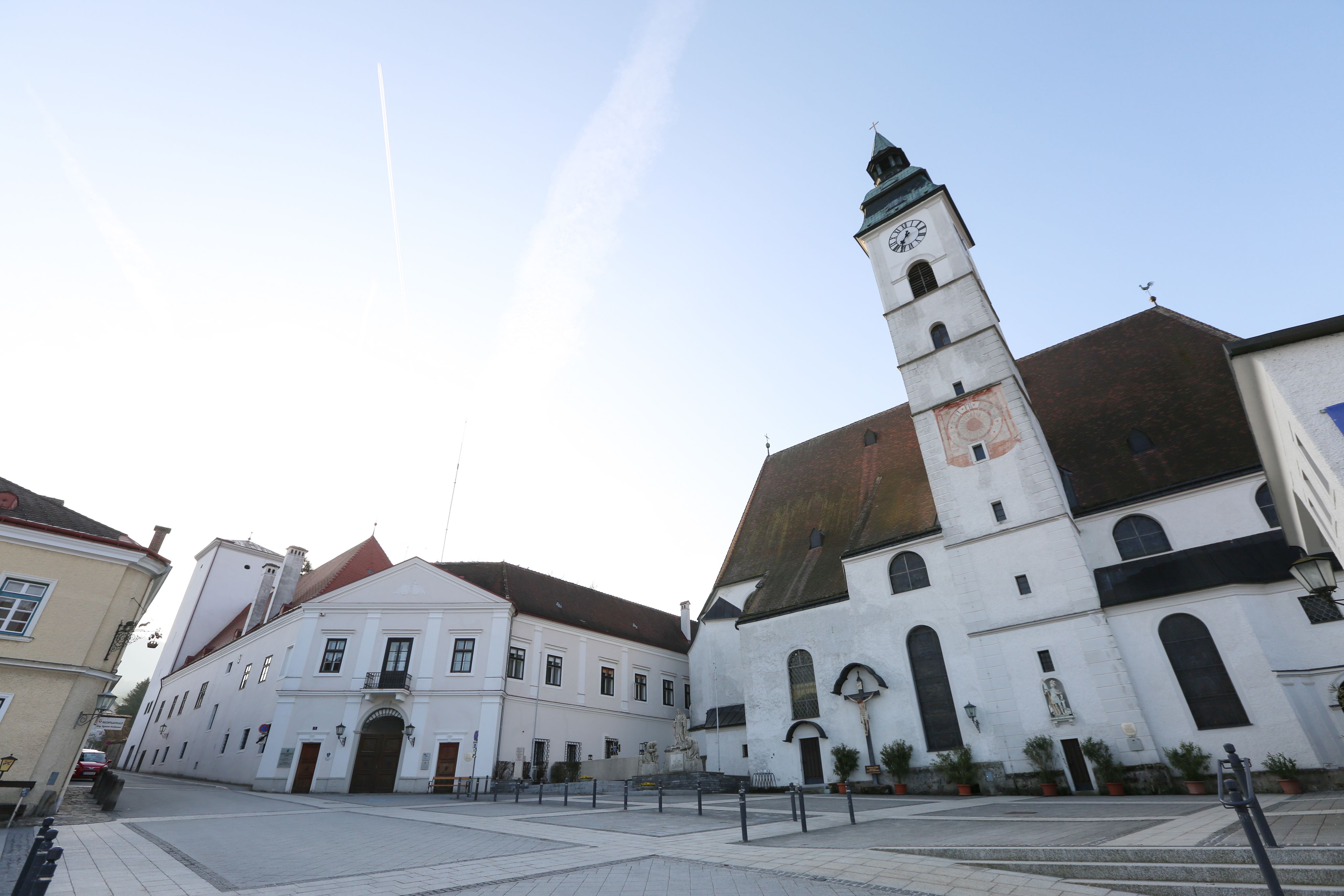 Farní kostel s hodinovou věží a sousedními budovami na prázdném náměstí.