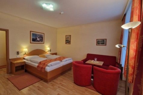 Hotelový pokoj s manželskou postelí, červenou pohovkou a křesly, dřevěnou podlahou a nástěnnými malbami.