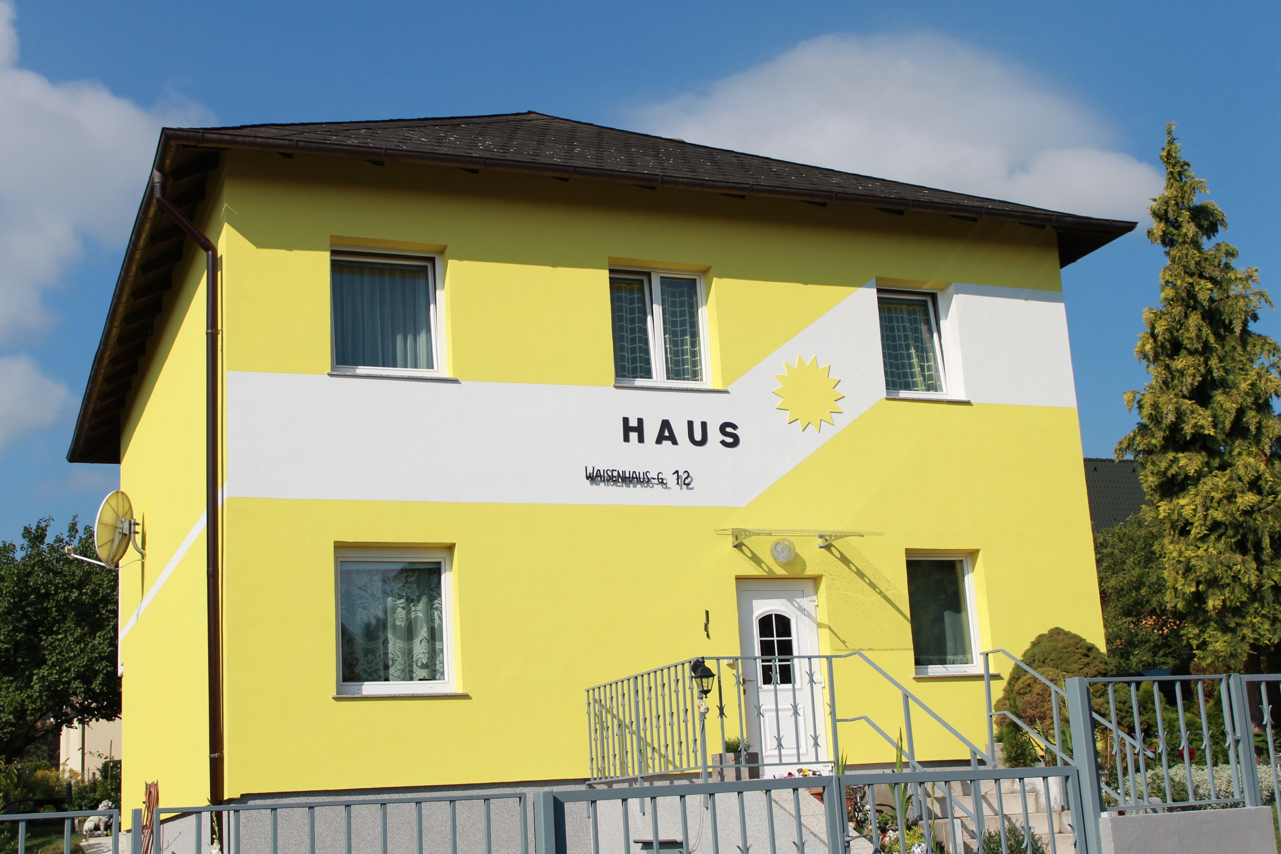 Žlutý dům s nápisem "Haus" a sluncem na fasádě.