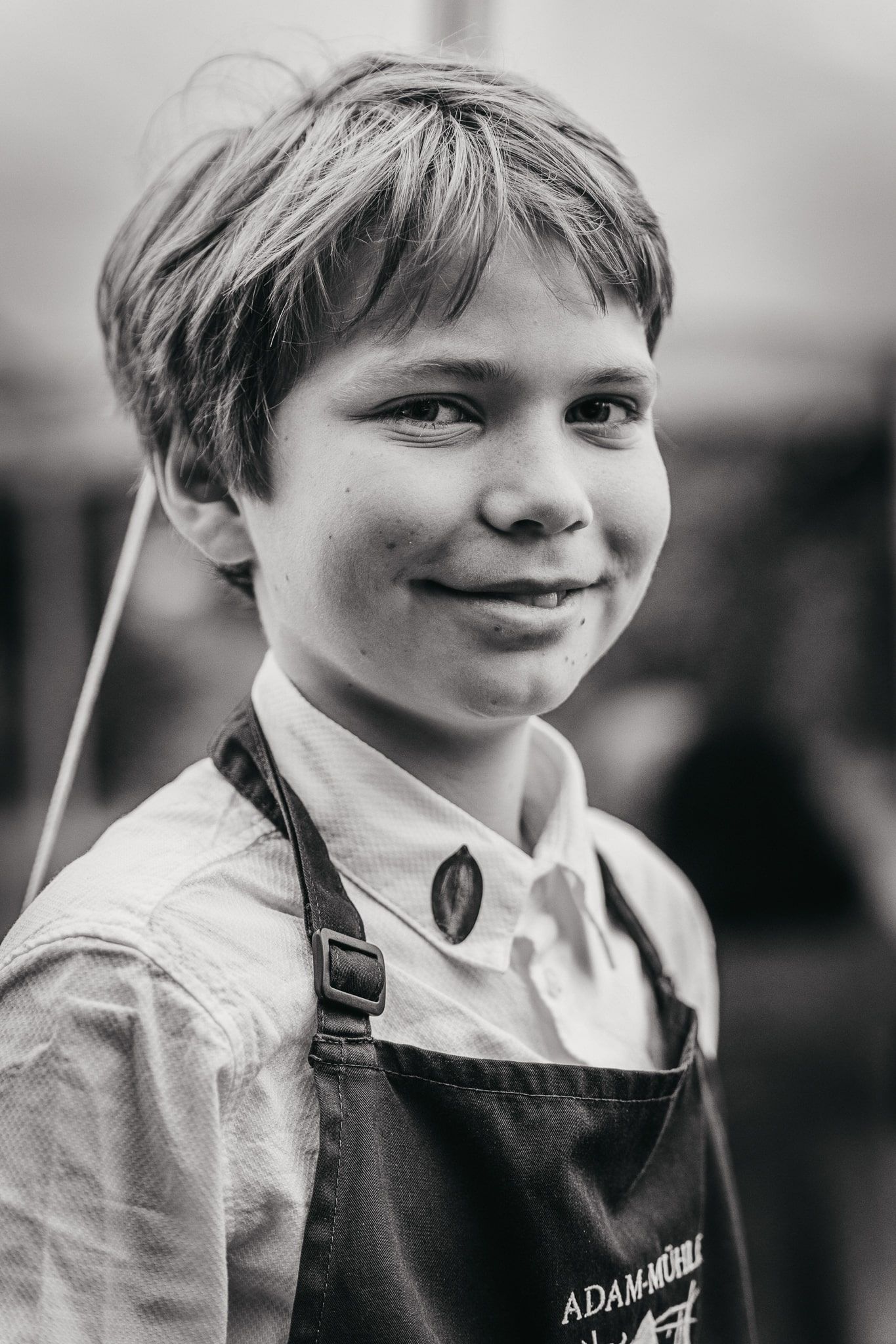 Chlapec v zástěře s nápisem "Adam-Mühle" se usmívá do kamery.