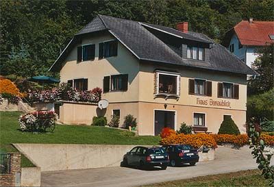 Dvoupatrový dům s nápisem "Haus Donaublick", obklopený květinami a stromy. Před domem parkují dvě auta.