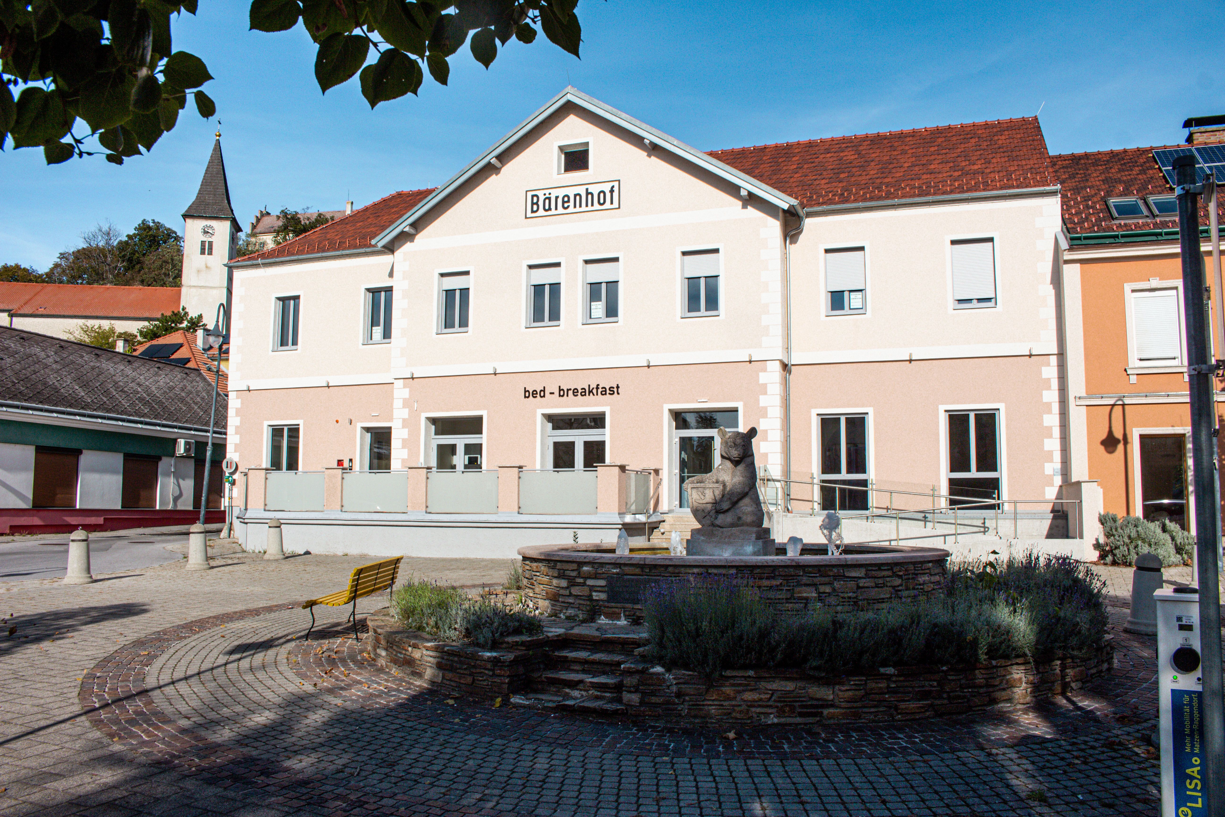 Budova s nápisem "Bärenhof" a "bed - breakfast" ve slunném prostředí s fontánou v popředí.