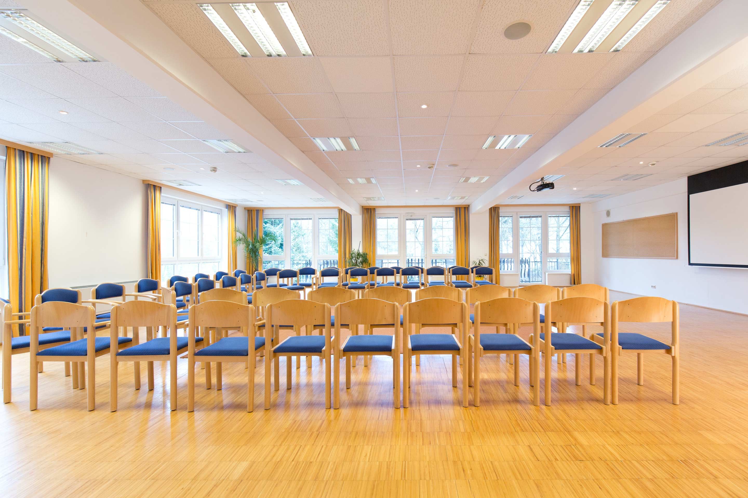 Světlá seminární místnost s řadami židlí, velkými okny a projektorem na stropě.