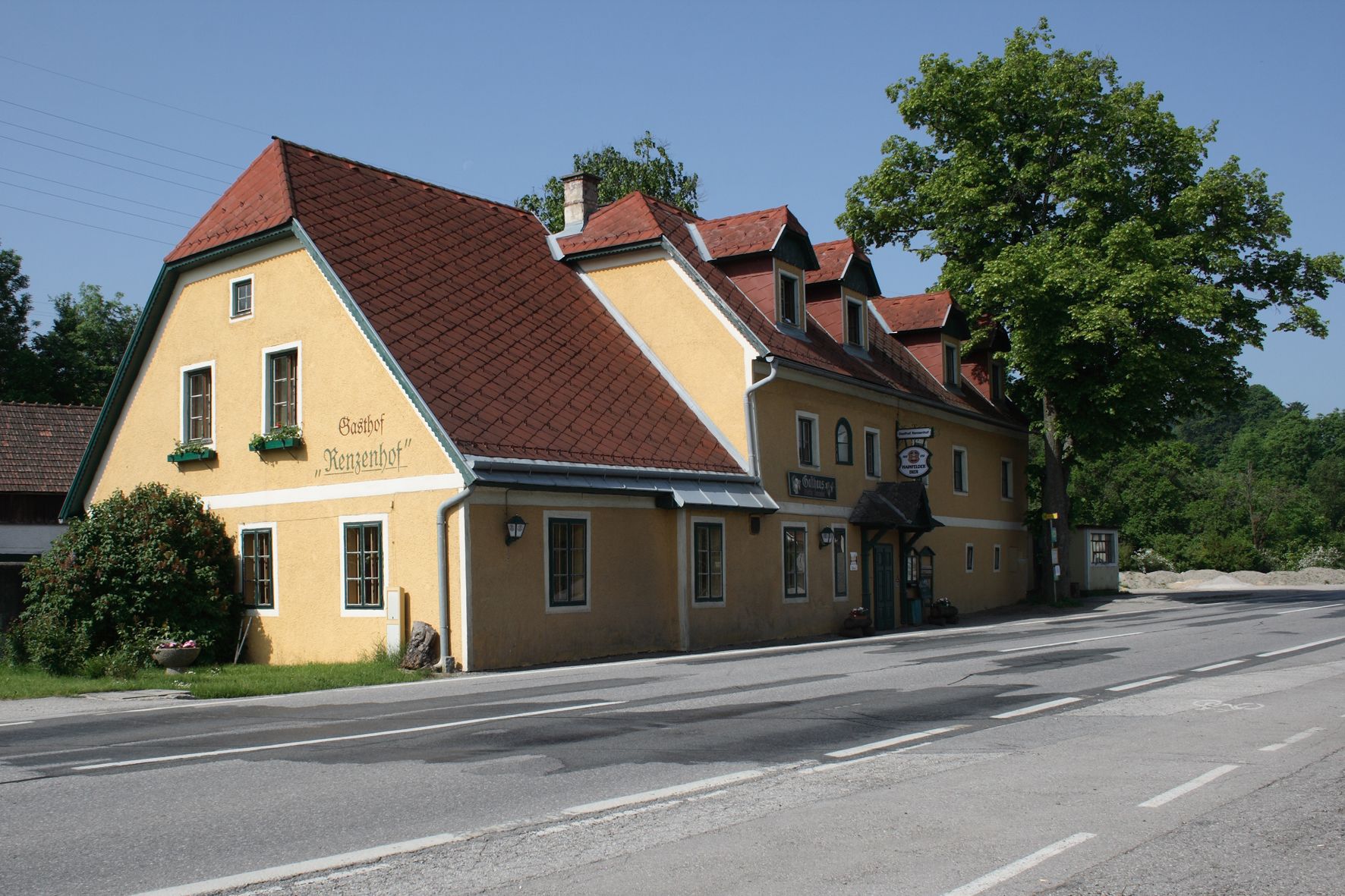 Žlutý hostinec s červenou střechou na silnici, obklopený stromy.