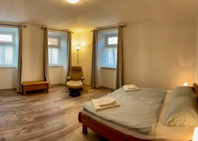 Jednoduchá ložnice s manželskou postelí, křeslem, nočním stolkem a třemi okny se závěsy.