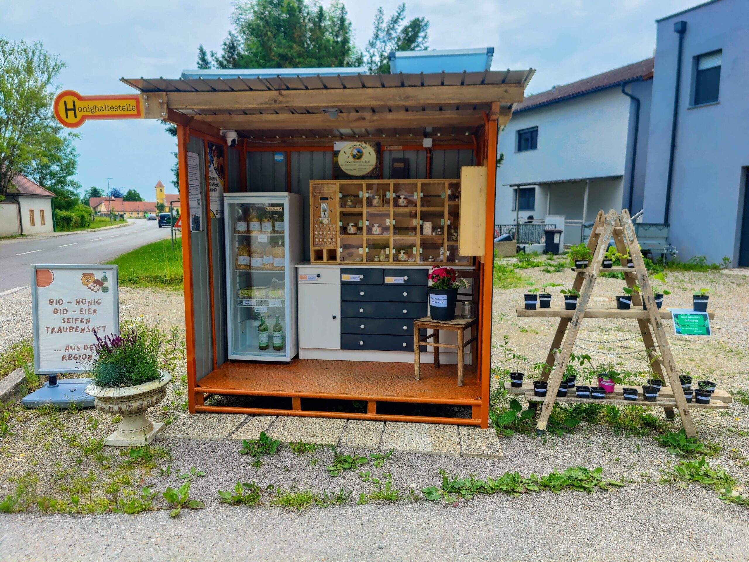 Malý stánek u silnice s bioprodukty, jako je med, vejce a mýdla. Vedle něj jsou rostliny na dřevěném rámu.