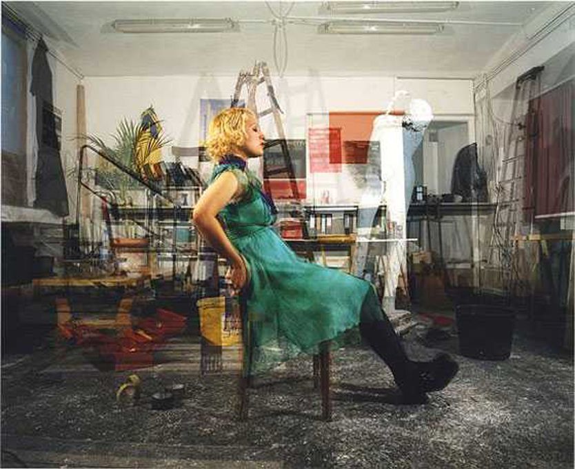 Žena v zelených šatech sedí na židli v přeplněné místnosti.