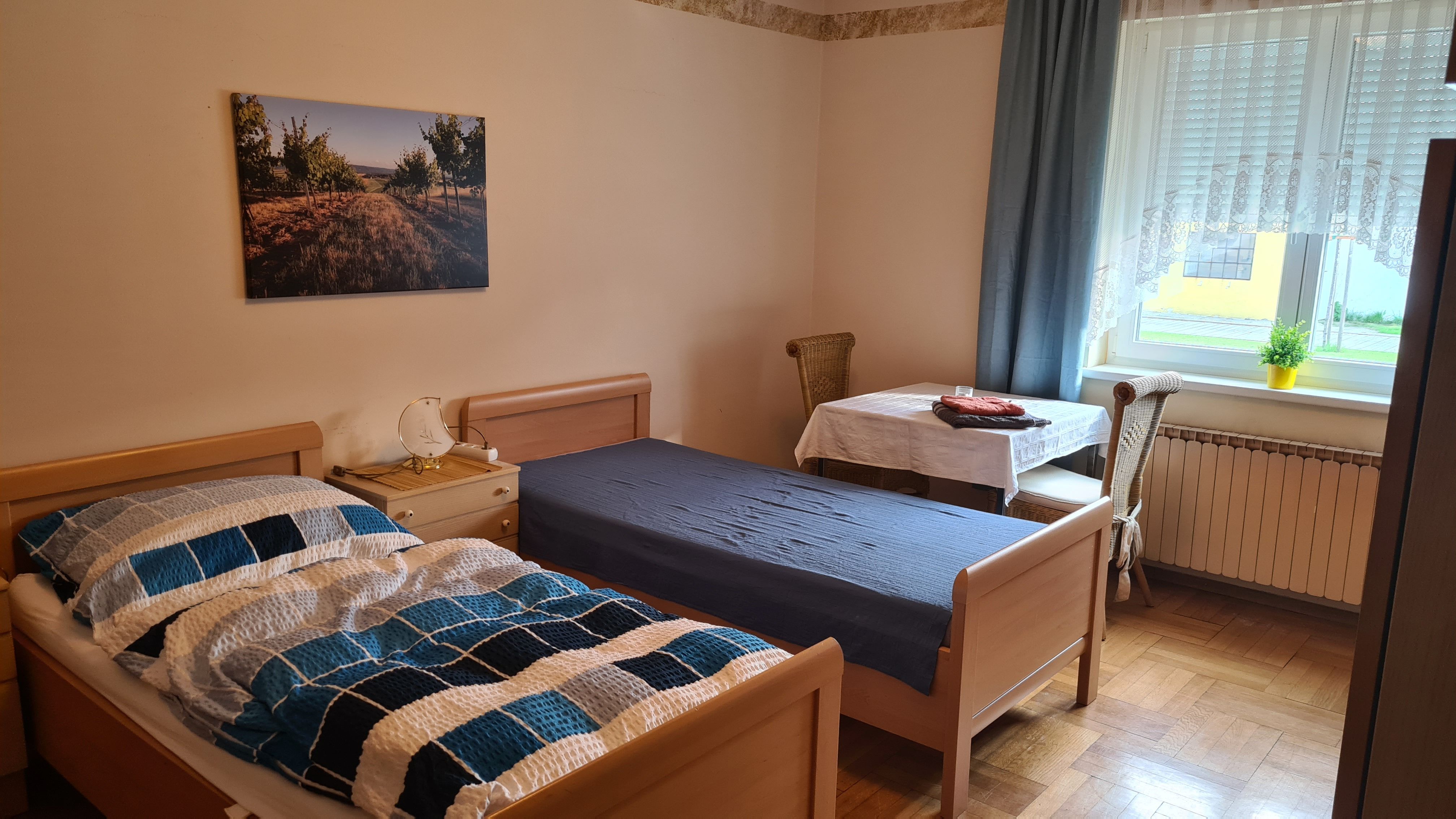 Jednoduše zařízená ložnice se dvěma samostatnými lůžky, stolem a židlí a oknem se závěsy.
