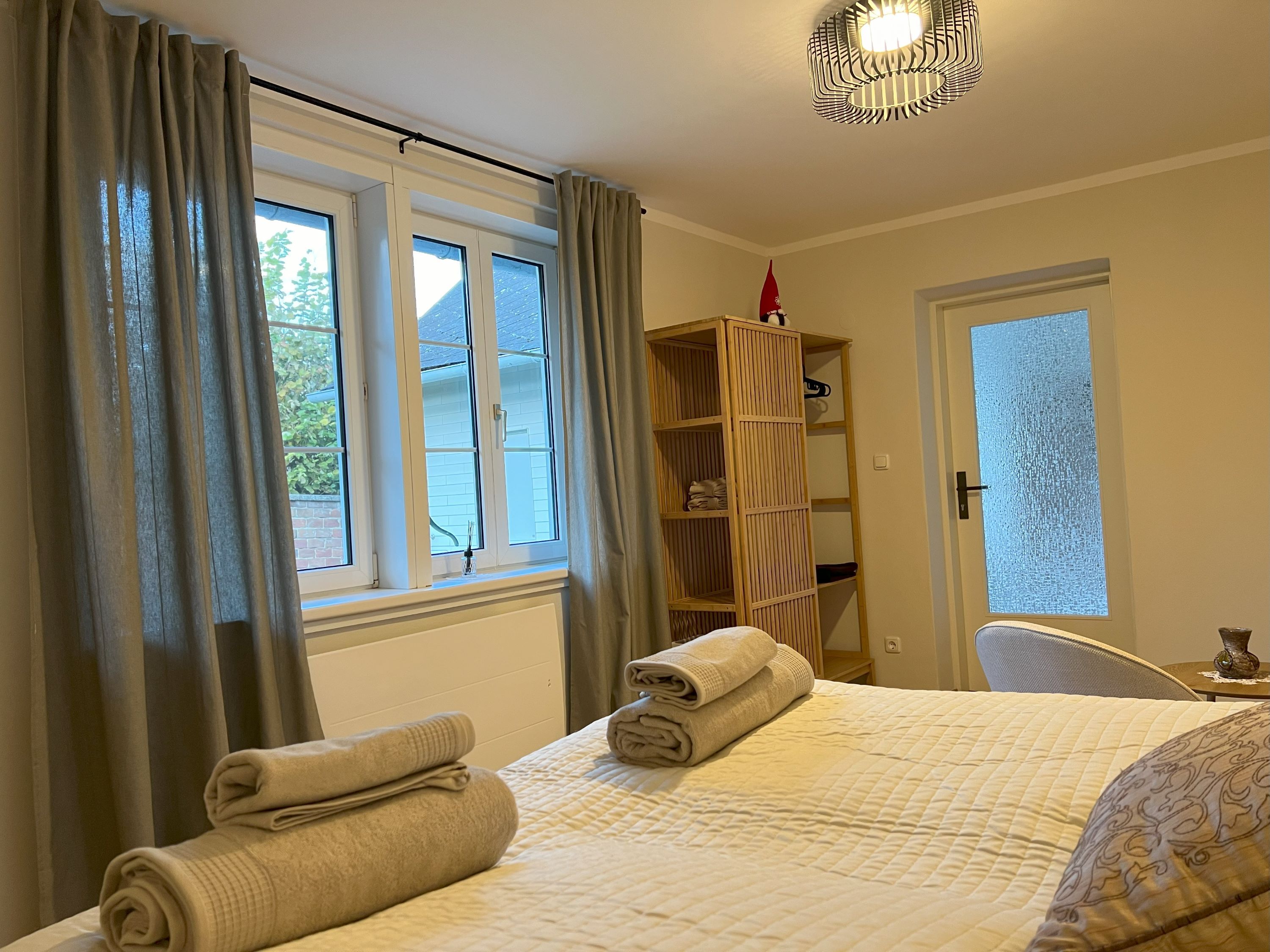 Útulná ložnice s postelí s ručníky, oknem se závěsy a poličkou s dekoracemi.