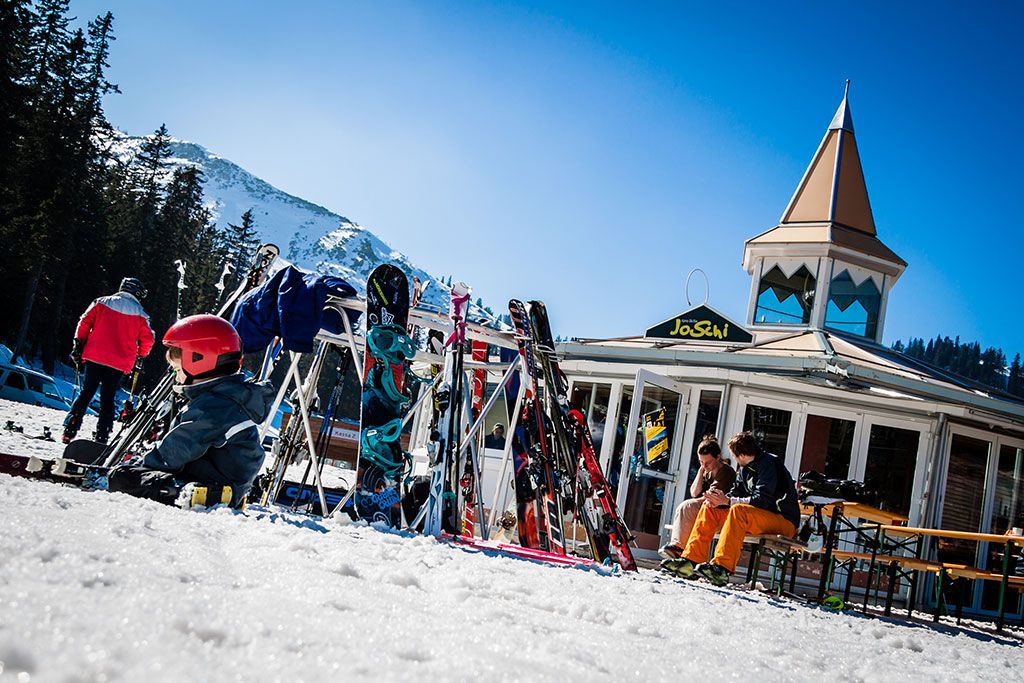 Lyžaři před barem JoSchi s lyžemi a snowboardy na sněhu.