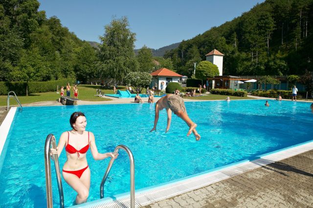 Venkovní bazén, kde se lidé koupou a relaxují. Žena vystupuje z vody, zatímco muž skáče do bazénu. Obklopují ho stromy a kopce.