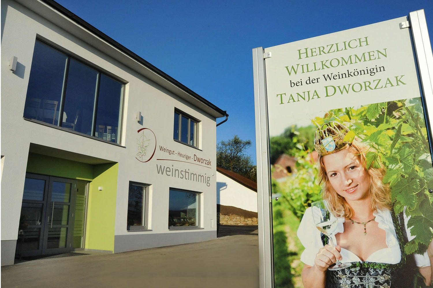 Moderní budova s nápisem zobrazujícím ženu v tradičním kroji se sklenicí vína. Text: Vítejte u královny vína Tanji Dworzak.