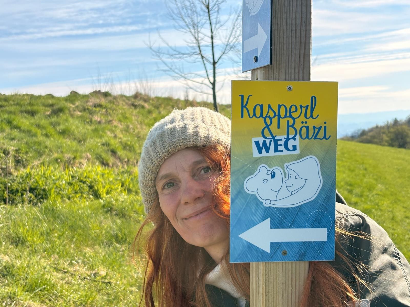Žena v čepici stojí vedle rozcestníku s nápisem „Kasperl & Bäzi Weg“ uprostřed zelené krajiny.