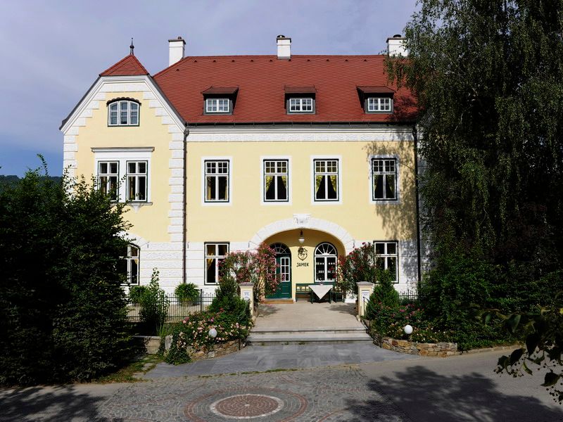 Žlutá budova s červenou střechou a zelenými dveřmi, obklopená stromy a rostlinami.