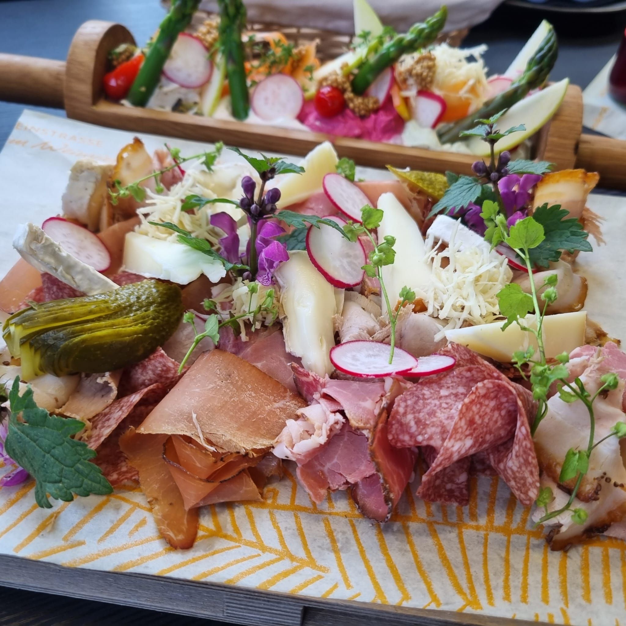 Talíř Heurigen s různými druhy uzenin a sýrů, ředkvičkami, okurkami a bylinkami podávaný na dřevěném prkénku.