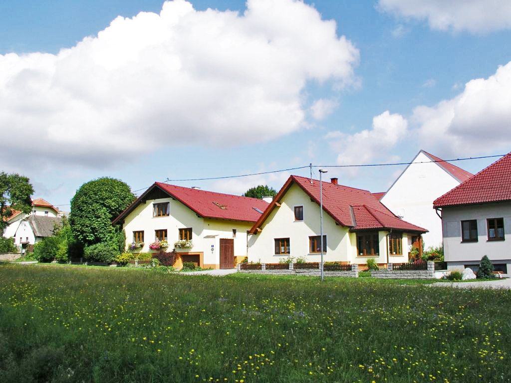 Ekologická farma s několika žlutými budovami s červenými střechami, obklopená zelenými loukami a stromy pod modrou oblohou s mraky.