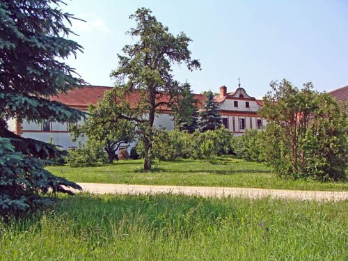 Historická budova s červenou střechou, obklopená stromy a zelenou loukou.