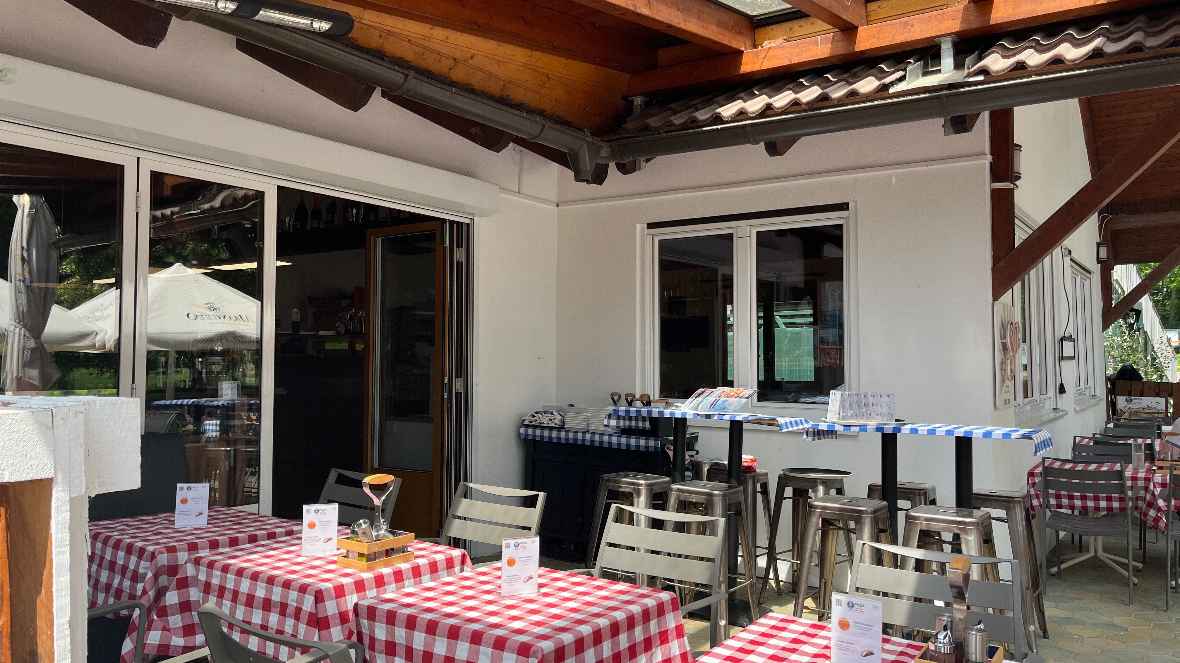 Vstupní prostor pizzerie se zahrádkou pro hosty. V popředí jsou prostřené stoly s červenými ubrusy.