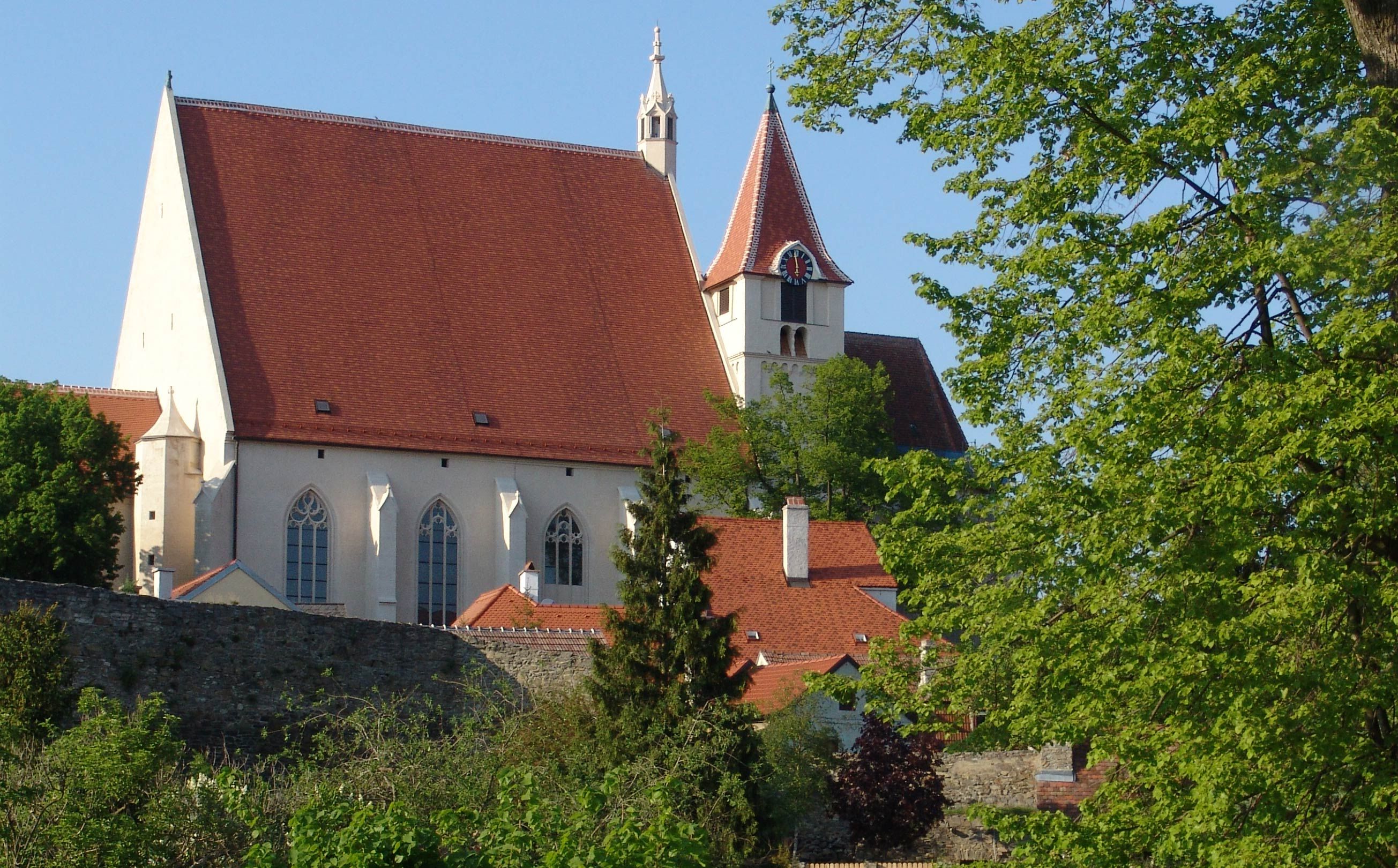Štěpána s červenou střechou a věží, obklopený stromy a kamennou zdí.