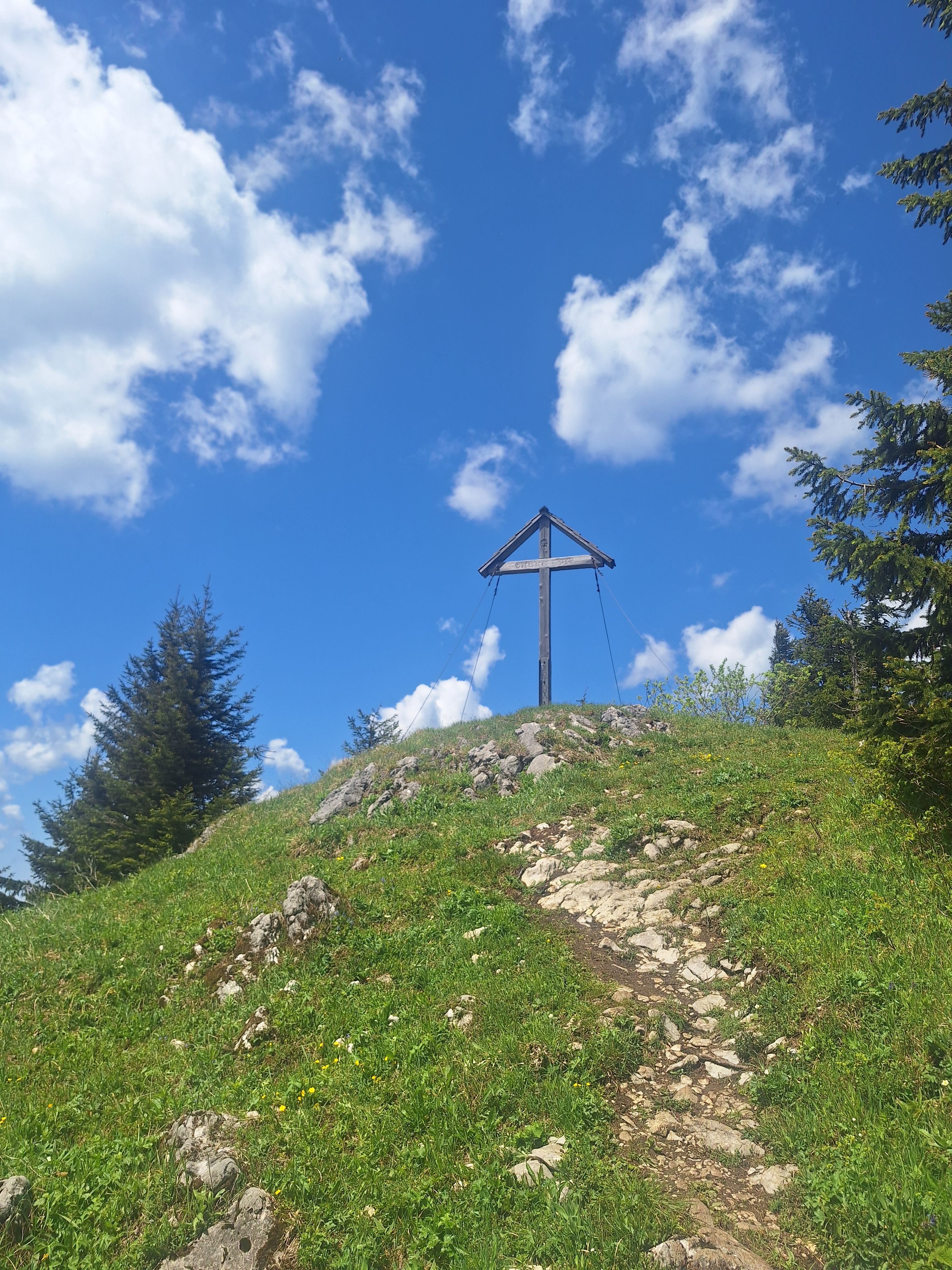 Vrcholový kříž na kopci s modrou oblohou a mraky.